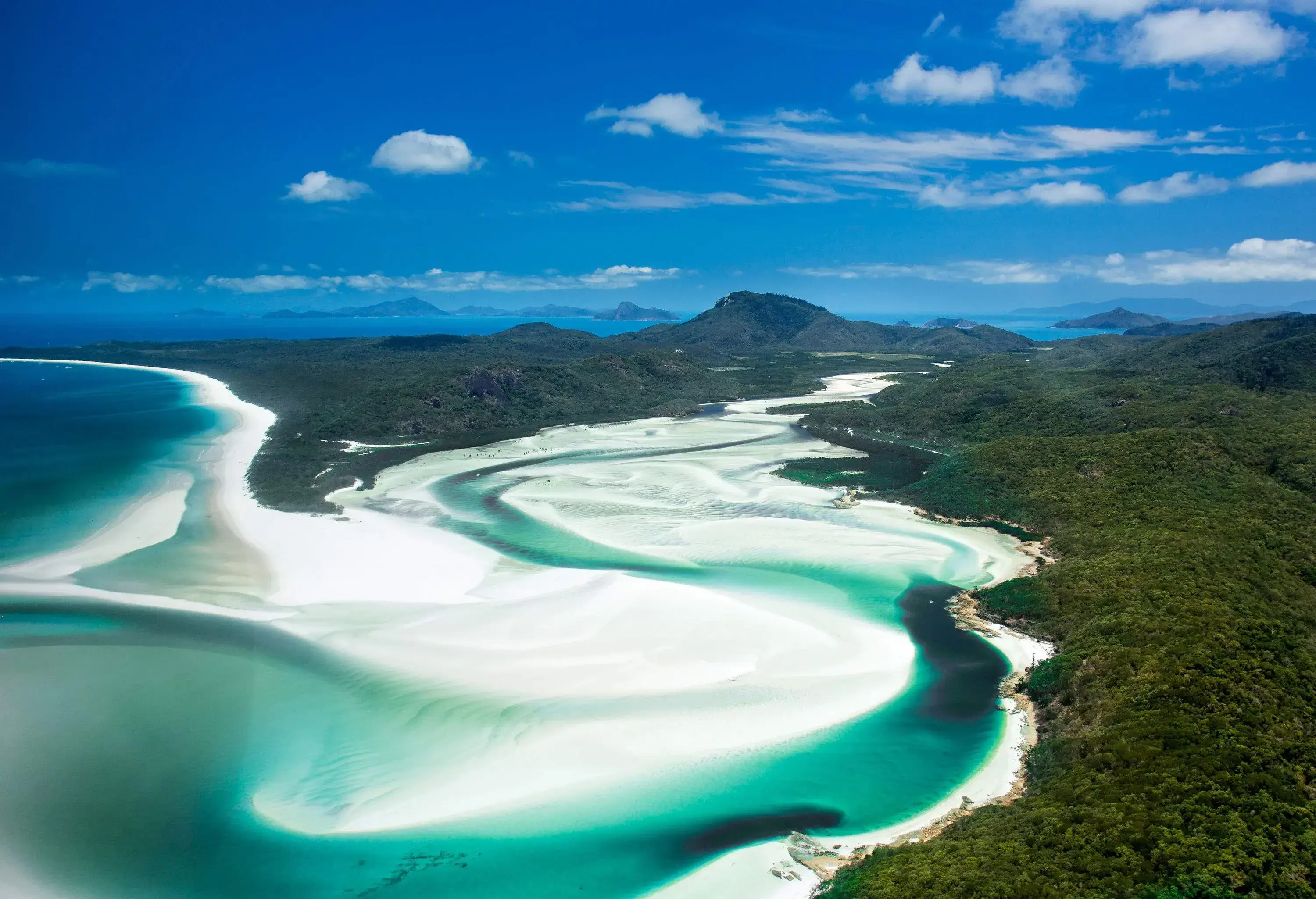 A beautiful blend of greenish blue ocean water and white sand snaking through wooded hills.