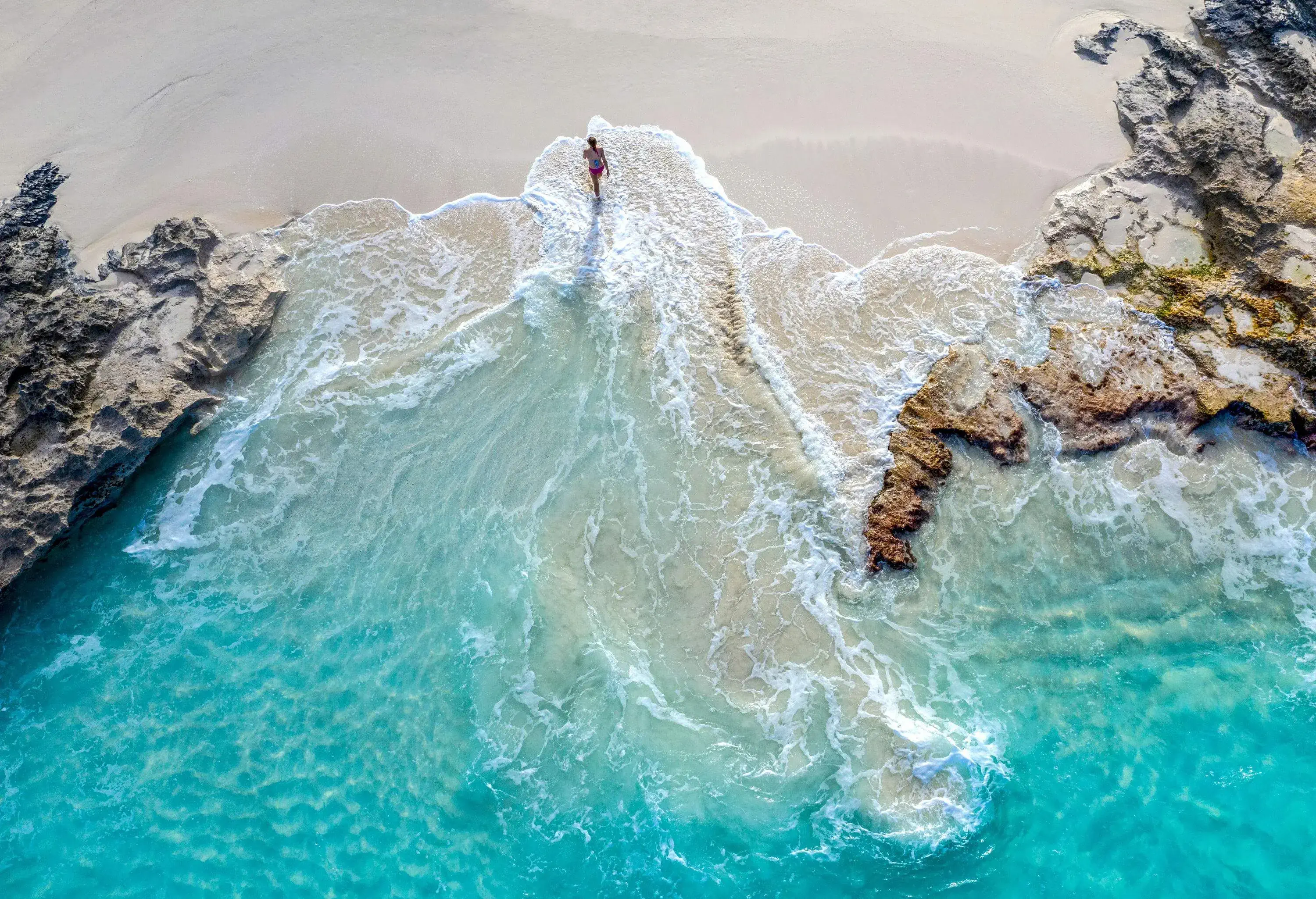 A person walks away from a beach with turquoise waves and rugged coast.