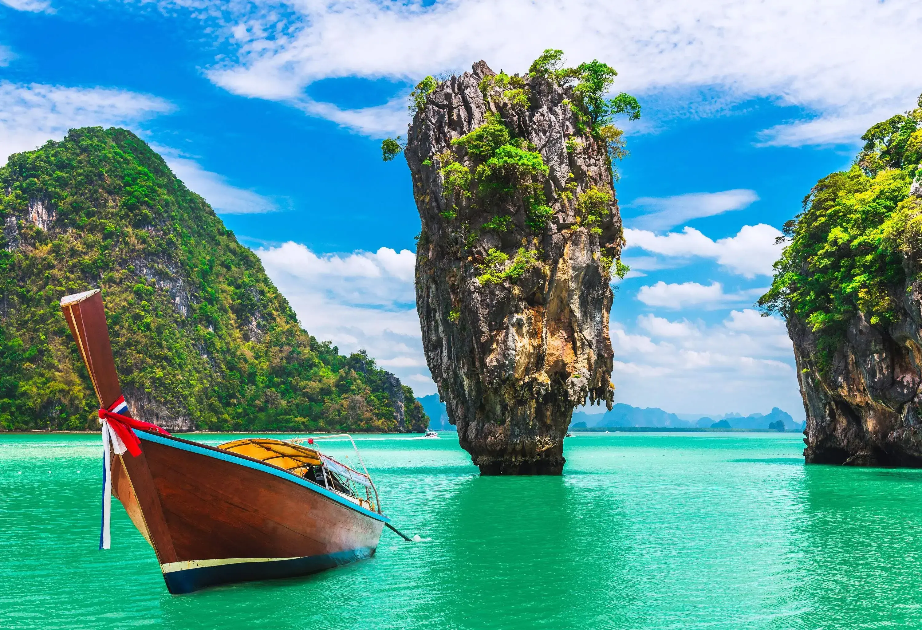 A wooden longtail boat on the tranquil turquoise sea with limestone cliffs shrouded in vegetation against the cloudy blue sky.