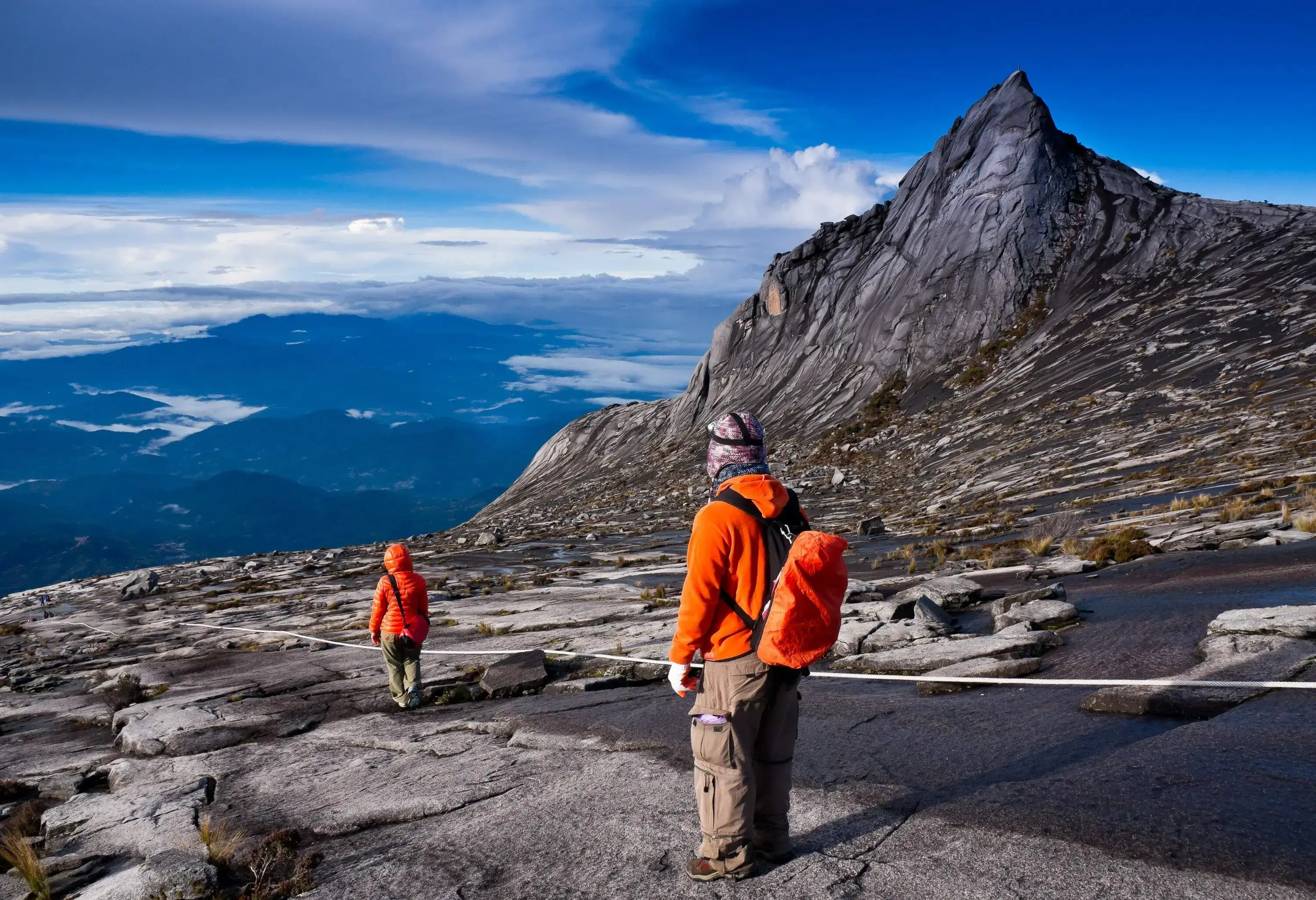 Two hikers in orange jackets traverse a trail with a rope beside a granite mountain peak.