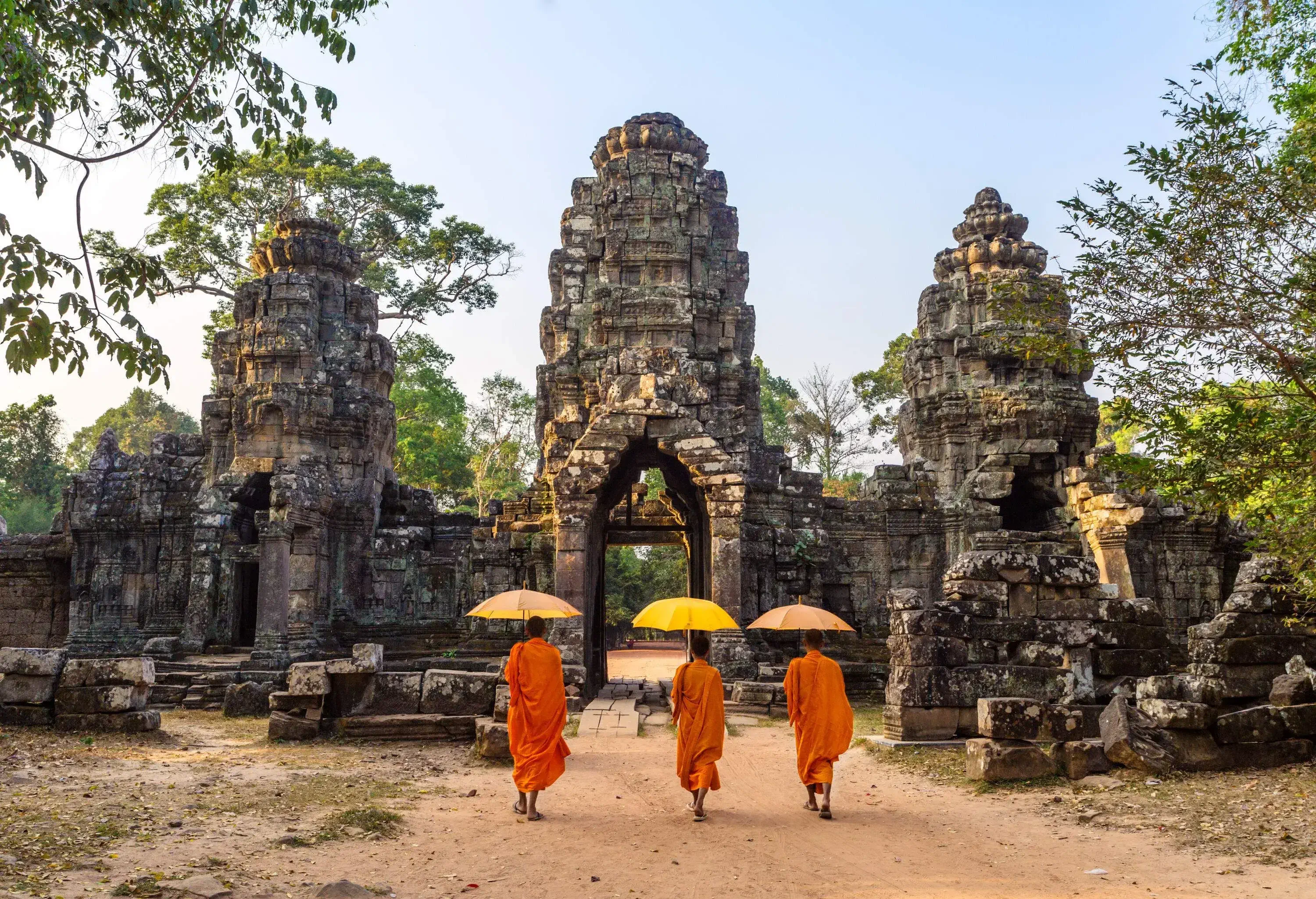 Three Buddhist monks, sheltered by their umbrellas, gracefully traverse the historic stone gate entrance of the majestic Angkor Wat temple complex.