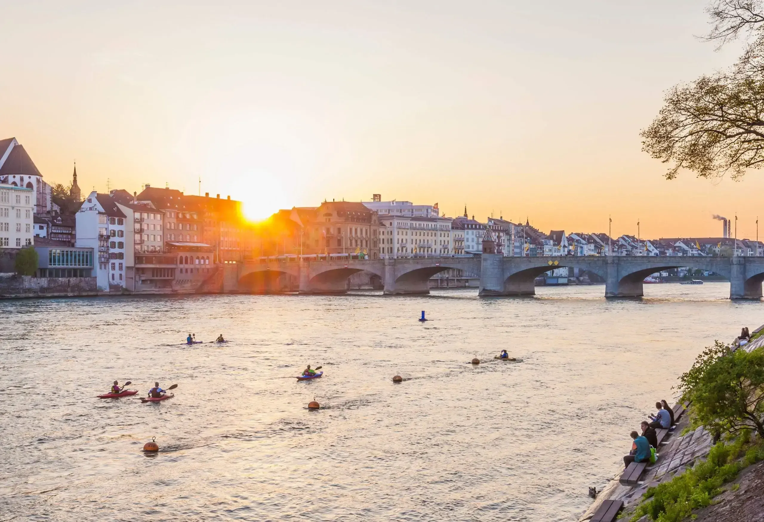 People on a promenade watch a group of kayakers paddling across a river towards an arched bridge that leads to a cluster of waterfront buildings.