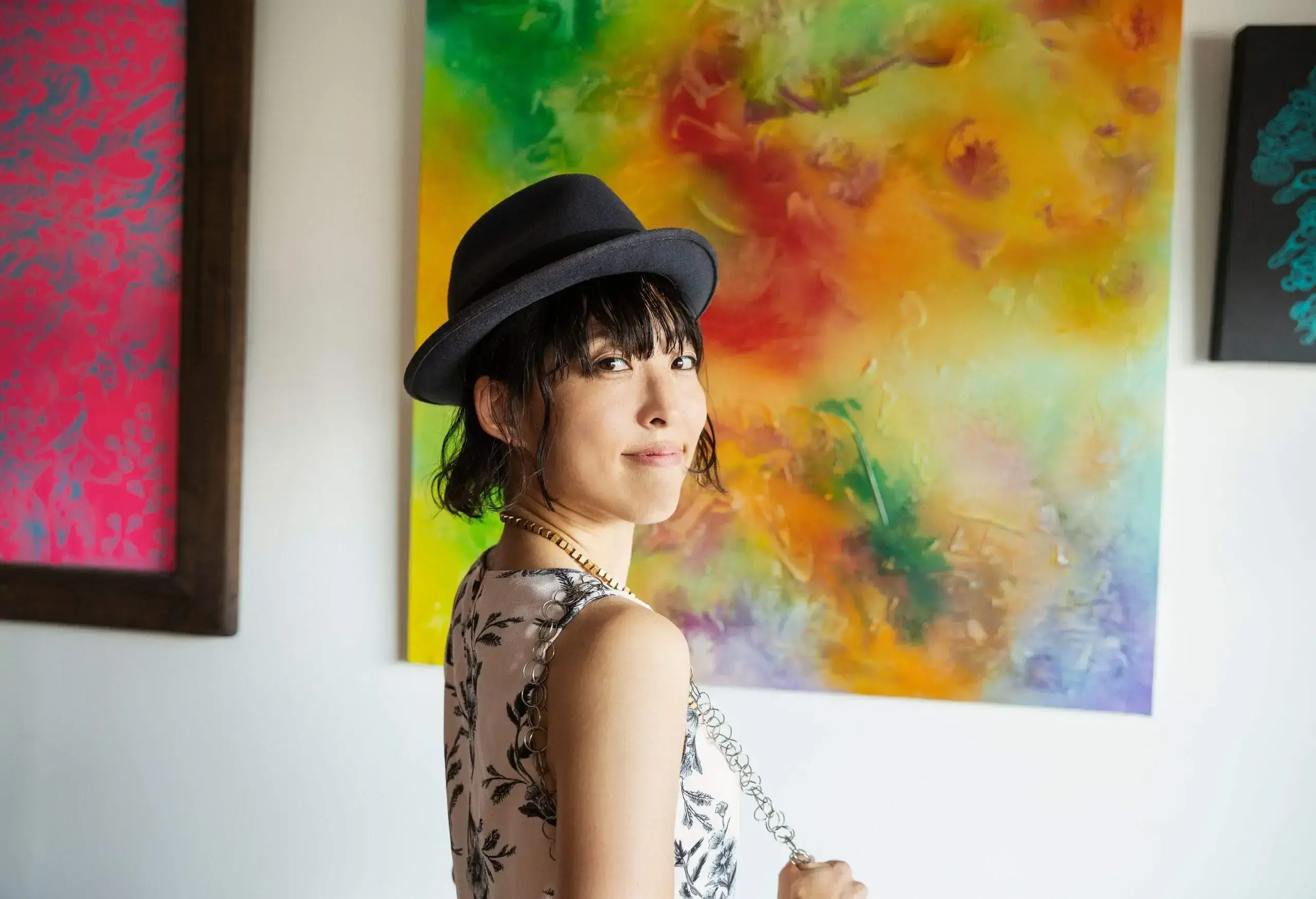 A young woman in a black hat stands in front of colourful paintings in a museum.