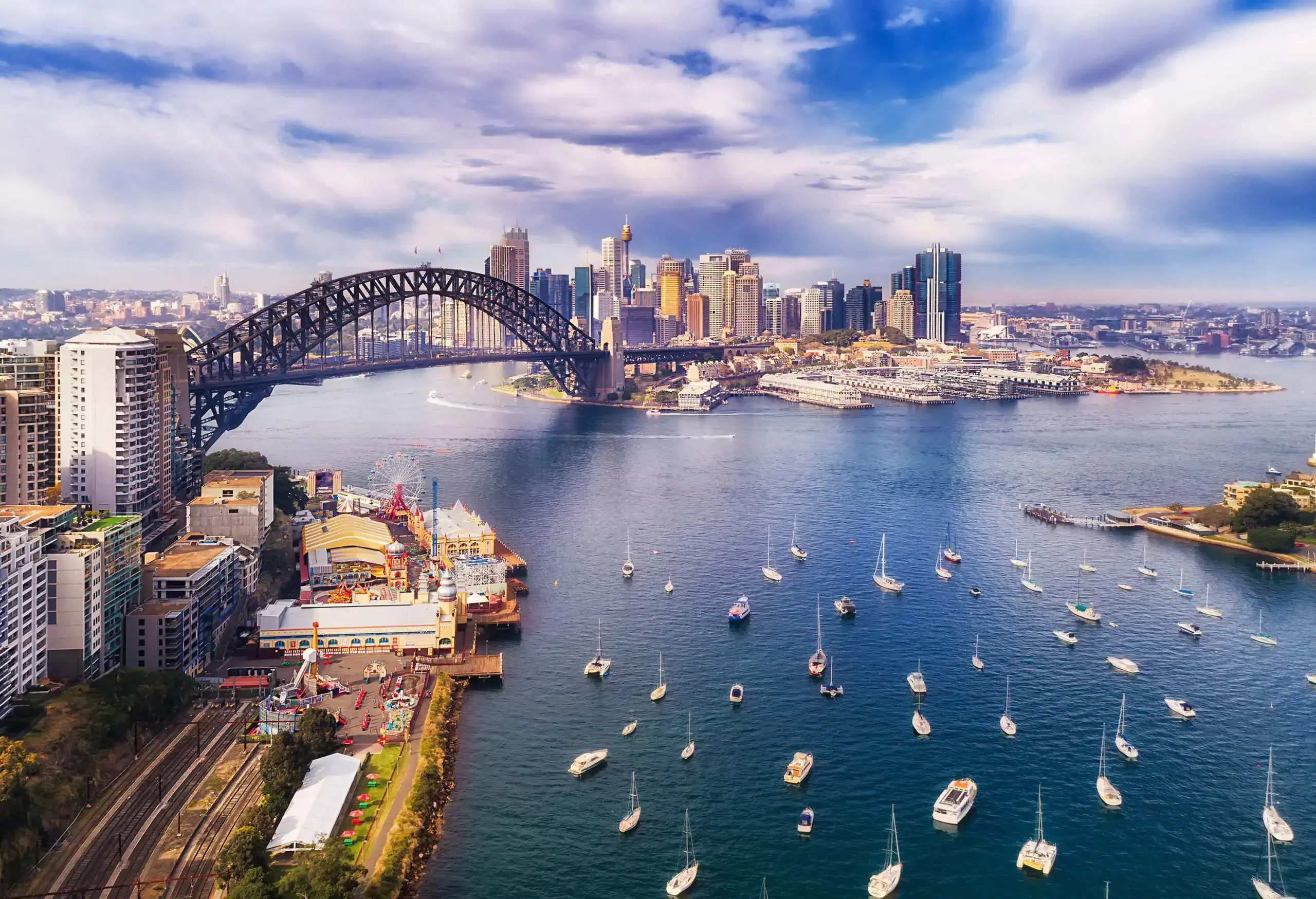 A steel arch bridge spans over the harbour connecting the modern coastal cities framed under the scenic cloudy sky.