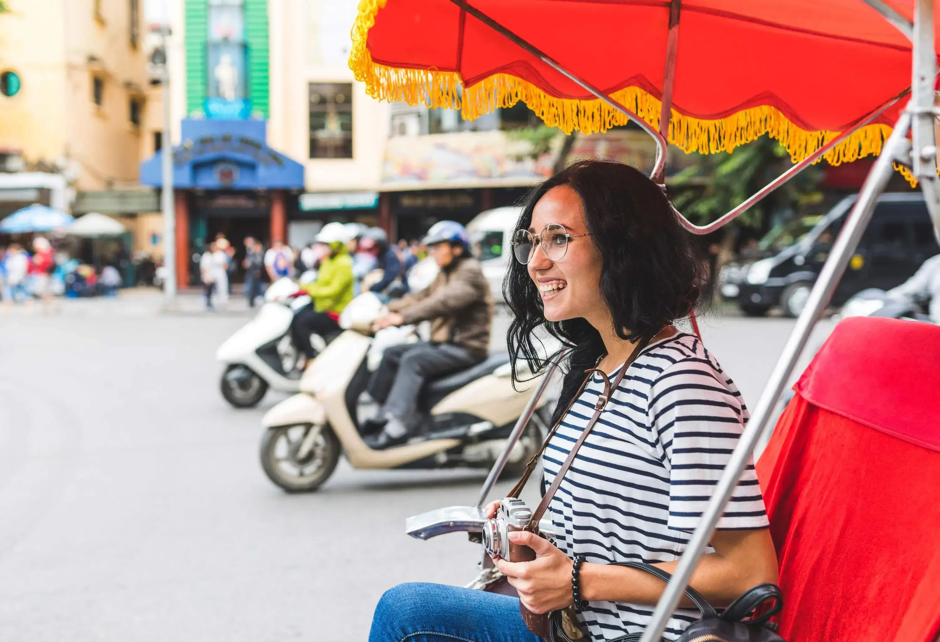 A woman in casual clothes smiles as she rides a rickshaw while holding a camera.