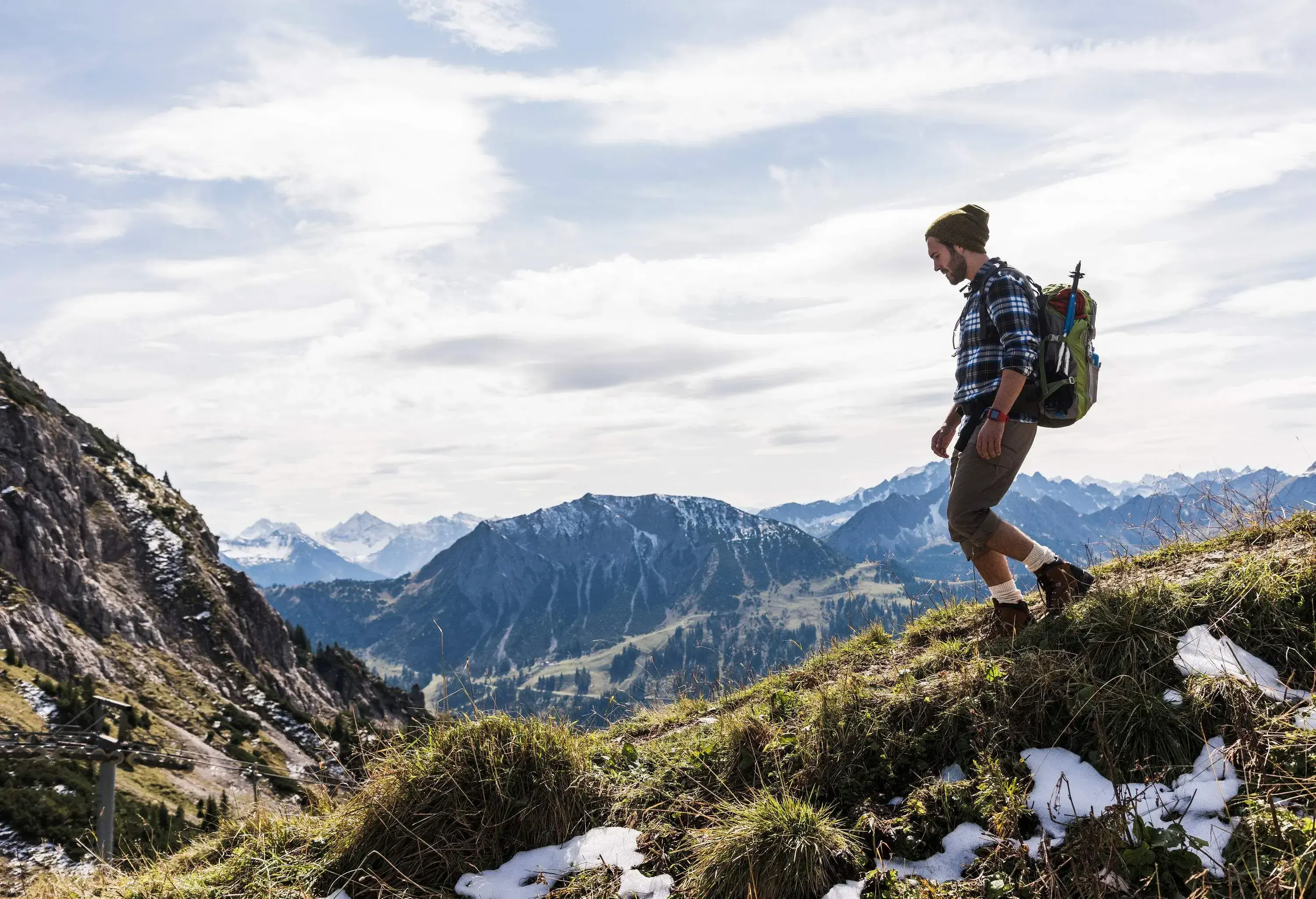 Hiker wearing a plaid shirt and carrying a bag descends a grassy slope in the mountains.