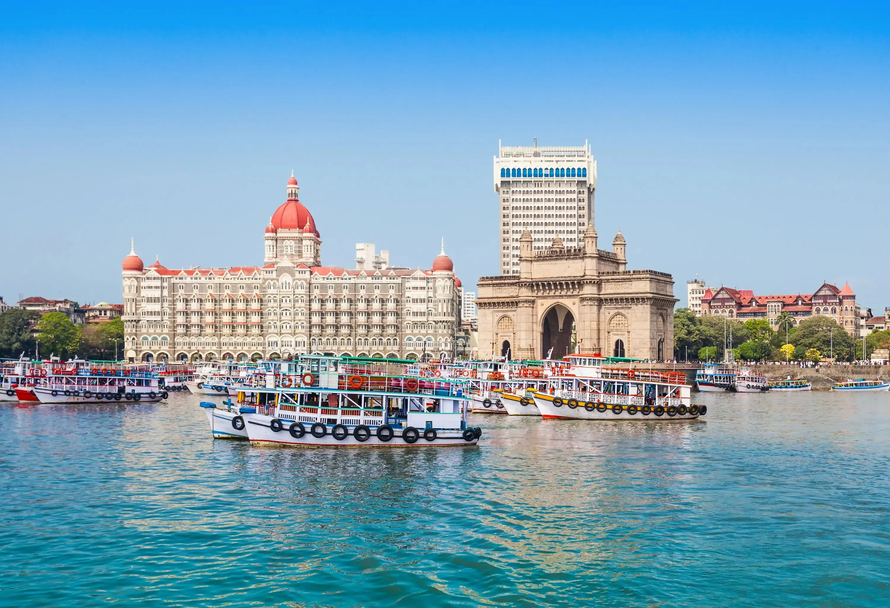 Colourful passenger boats moored on the harbour with a gigantic arch gateway monument next to the iconic Taj Mahal Palace.