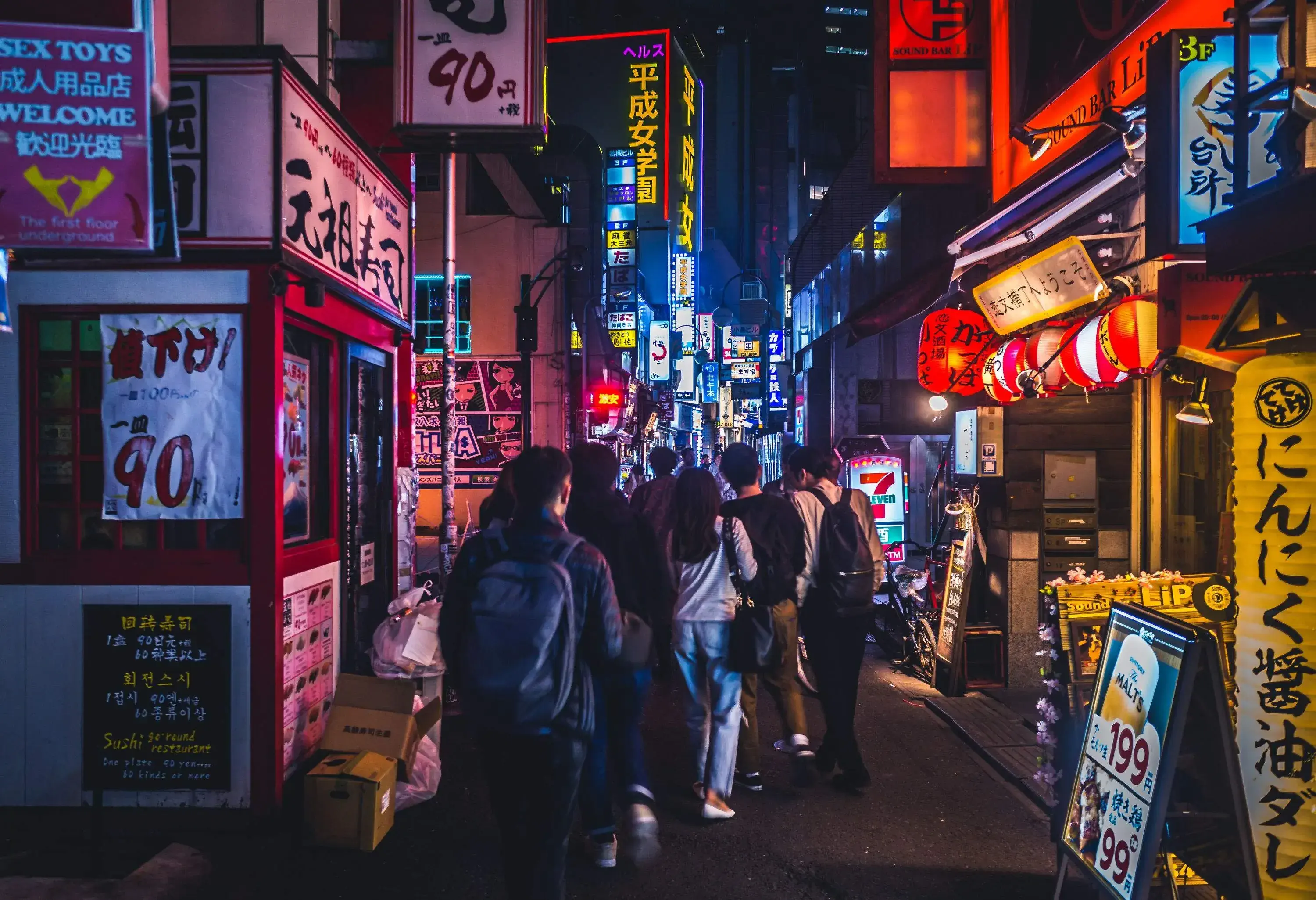 A night-time view of a crowded alley bordered by restaurants and bars with bright LED billboards.