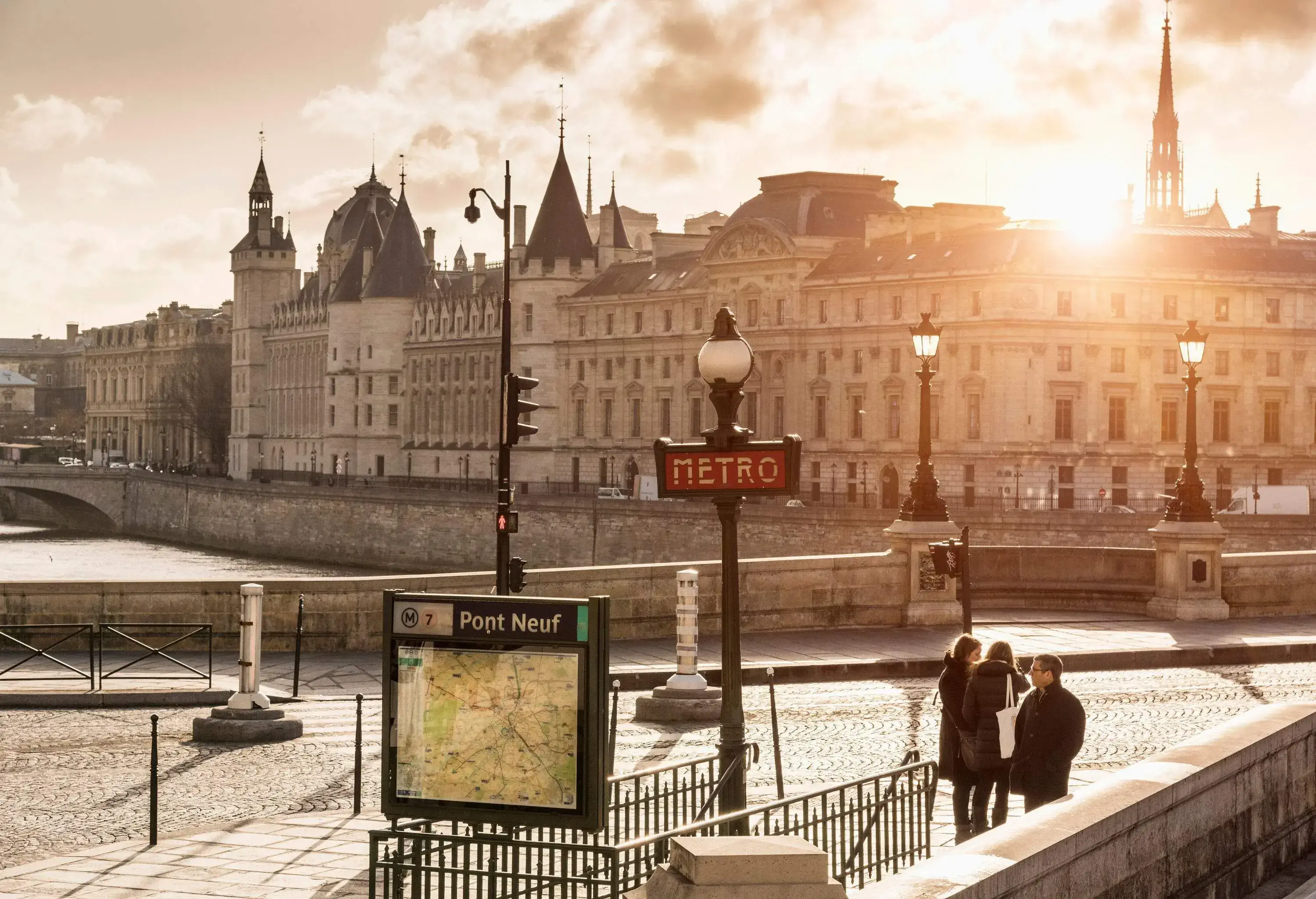 Three people standing along Pont Neuf as the sun blasts through the building rooftops.