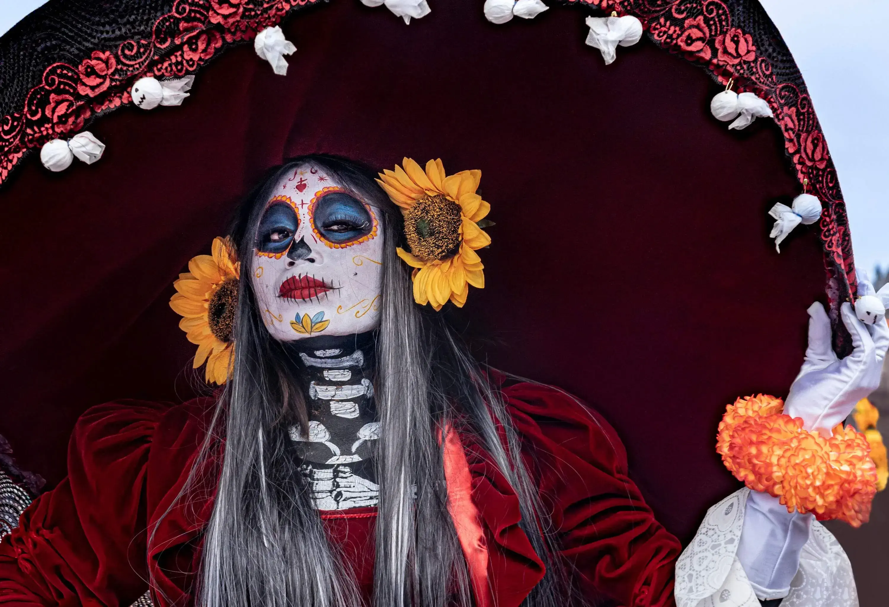A long-haired woman wearing a skull makeup and sunflowers behind her ears to complete the look of La Calavera Catrina.