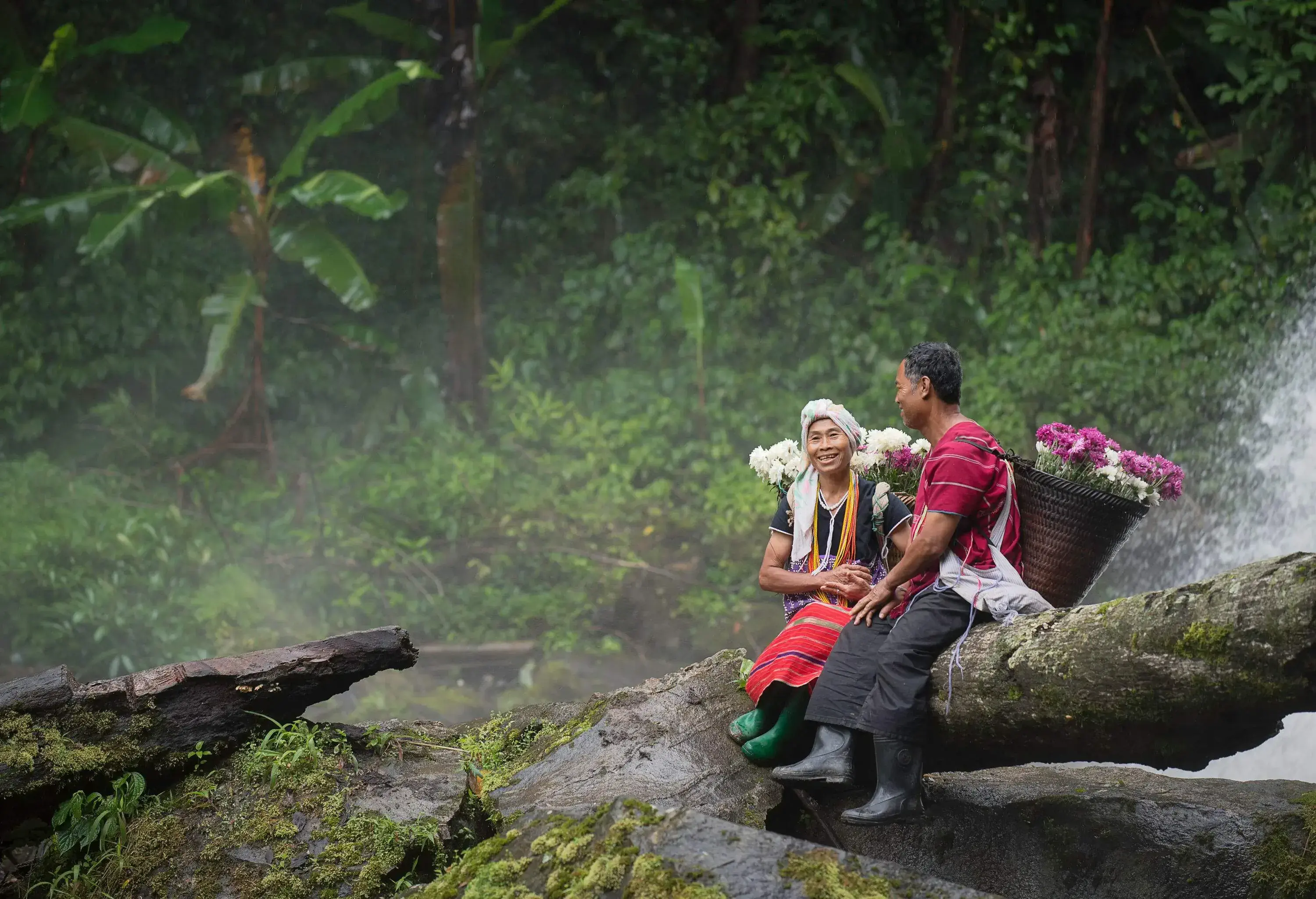 A couple in traditional attire carry baskets of flowers on their backs as they sit near a waterfall.