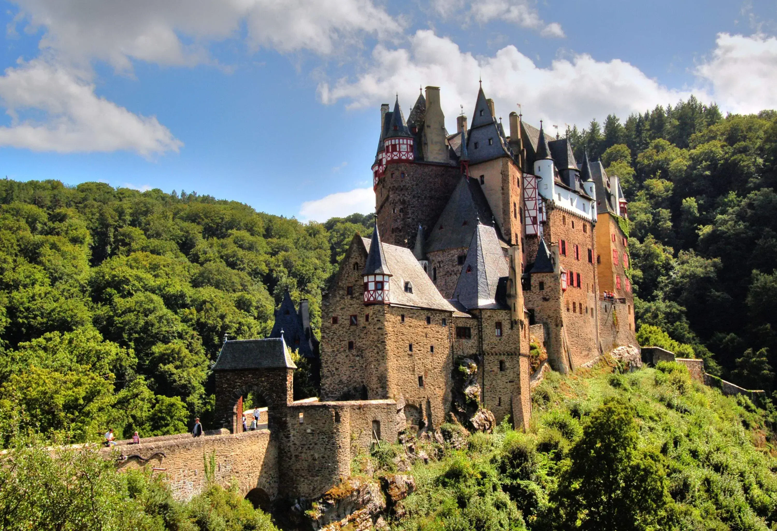 A medieval castle crowned on a hill surrounded by lush forested mountains.