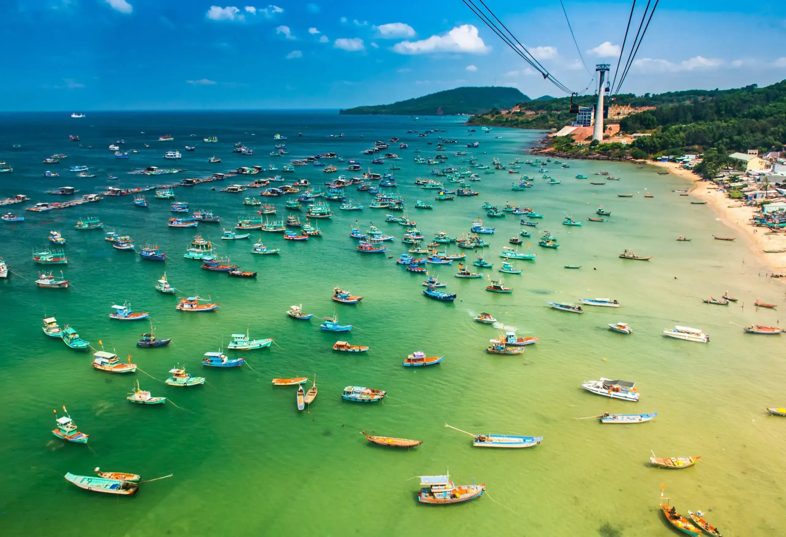 Cable cars travelling above the sea near the shore, occupied by numerous fishing boats docked on the shallow emerald green waters.