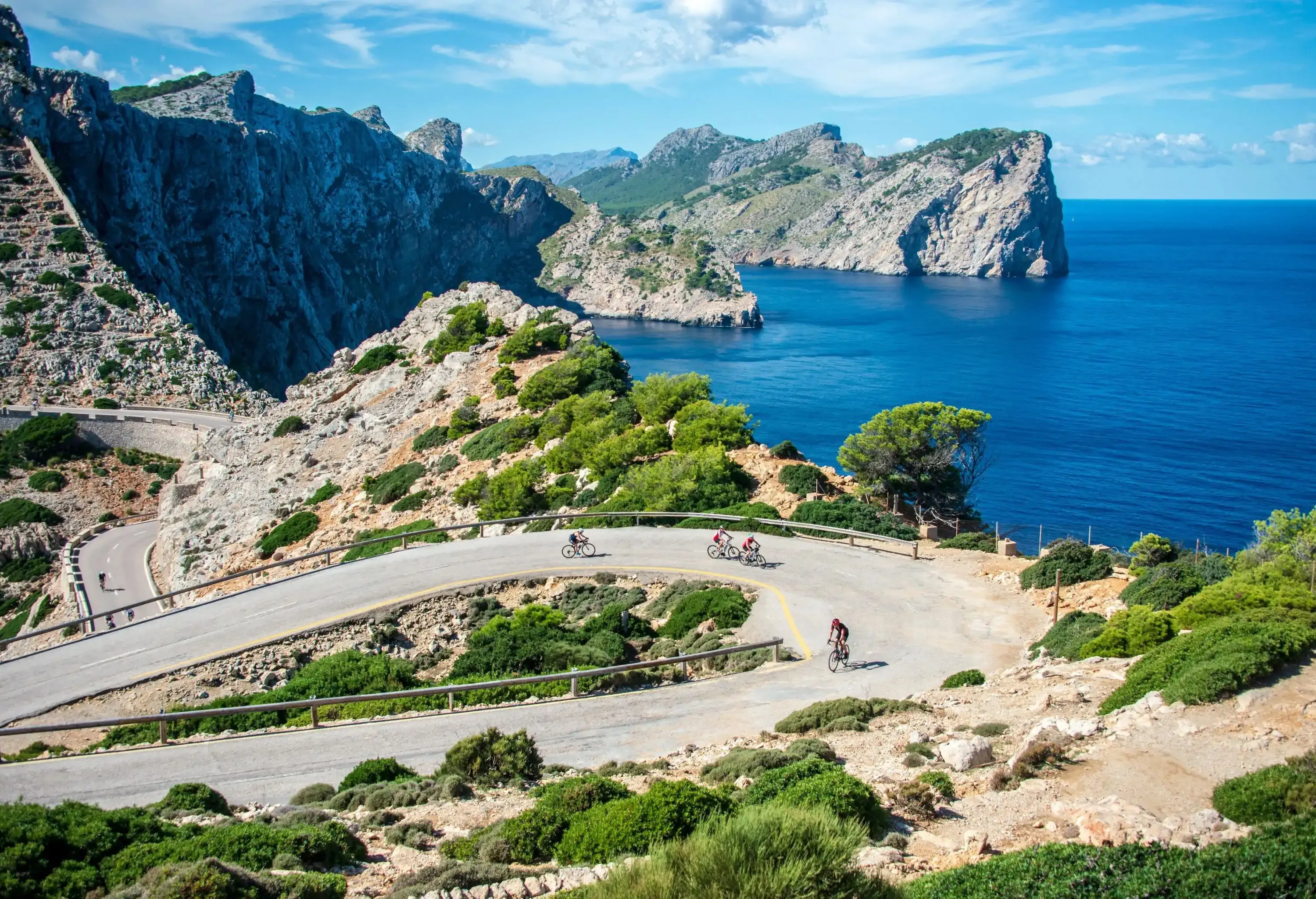 A group of cyclists travels on an uphill road on a mountainous area with views of rocky coastlines in the distance.