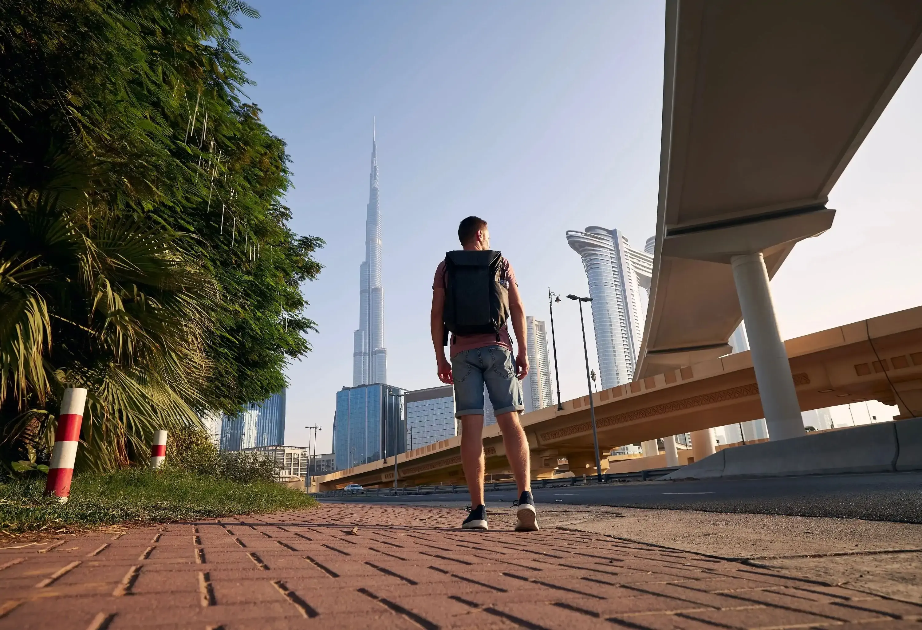 A man with his backpack stands in the middle of a promenade along a road under a skyway with a view of tall buildings.