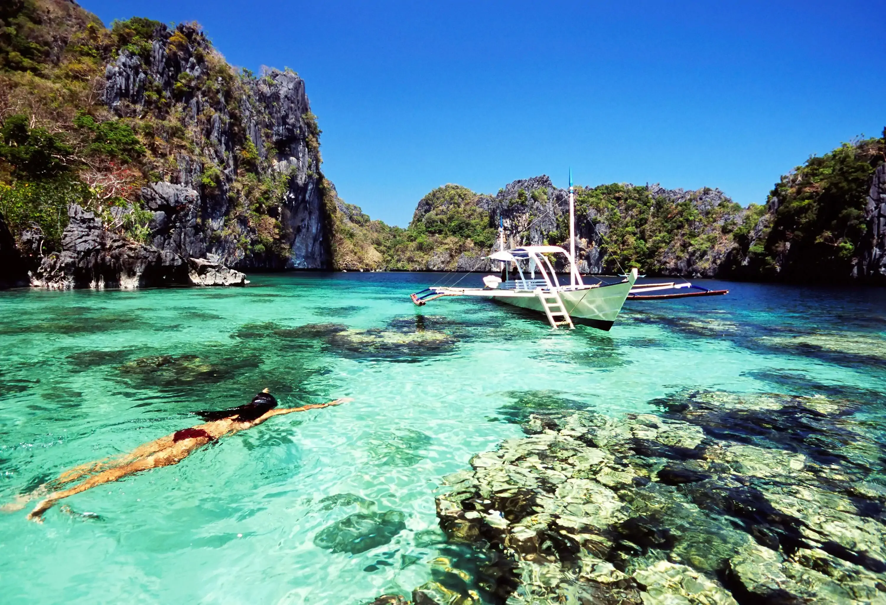 A woman swimming towards a docked boat in crystal clear calm water surrounded by rock mountains.