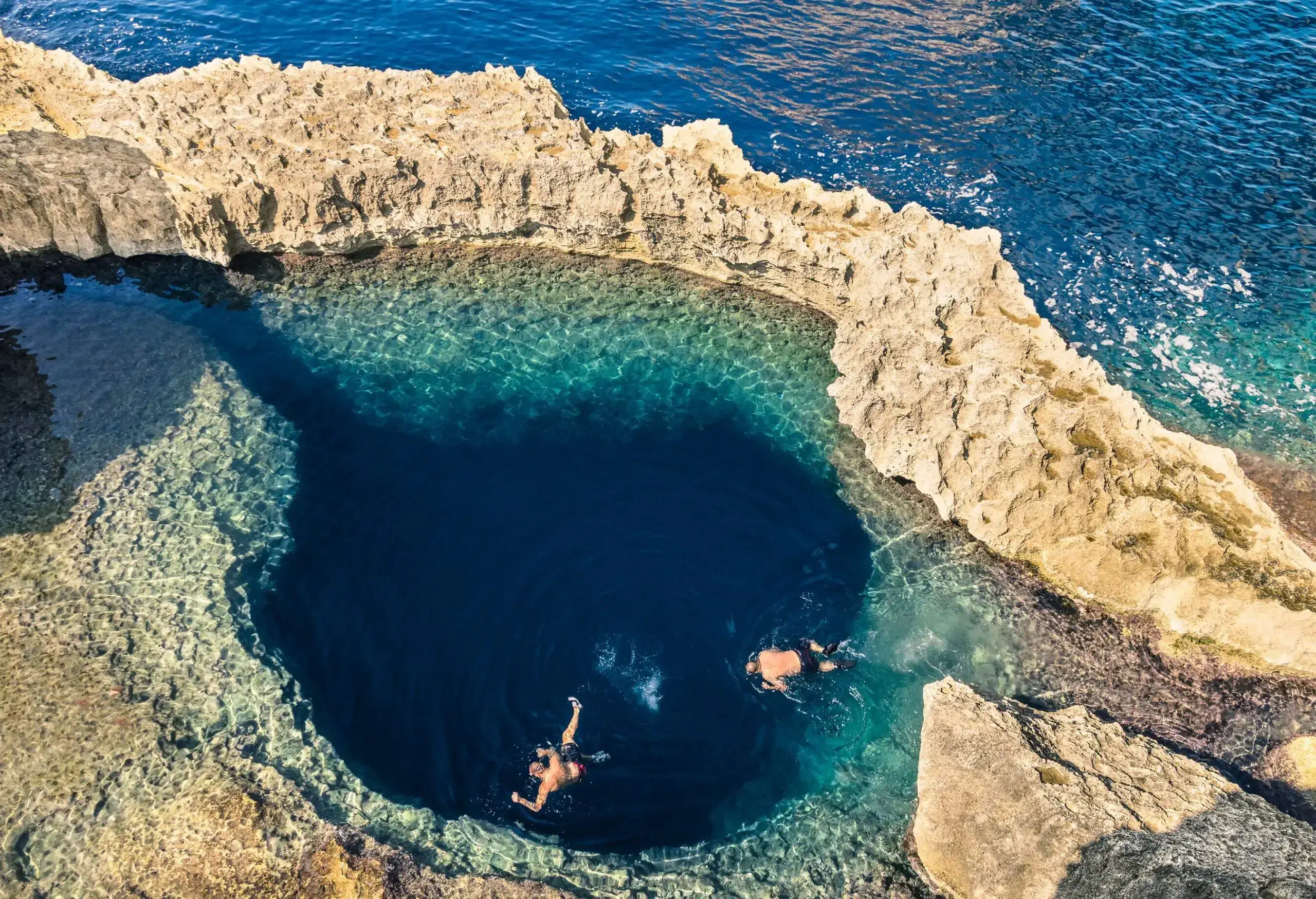 Two people are swimming in a large, deep blue hole next to the sea, which is encircled by a sedimentary rock formation.