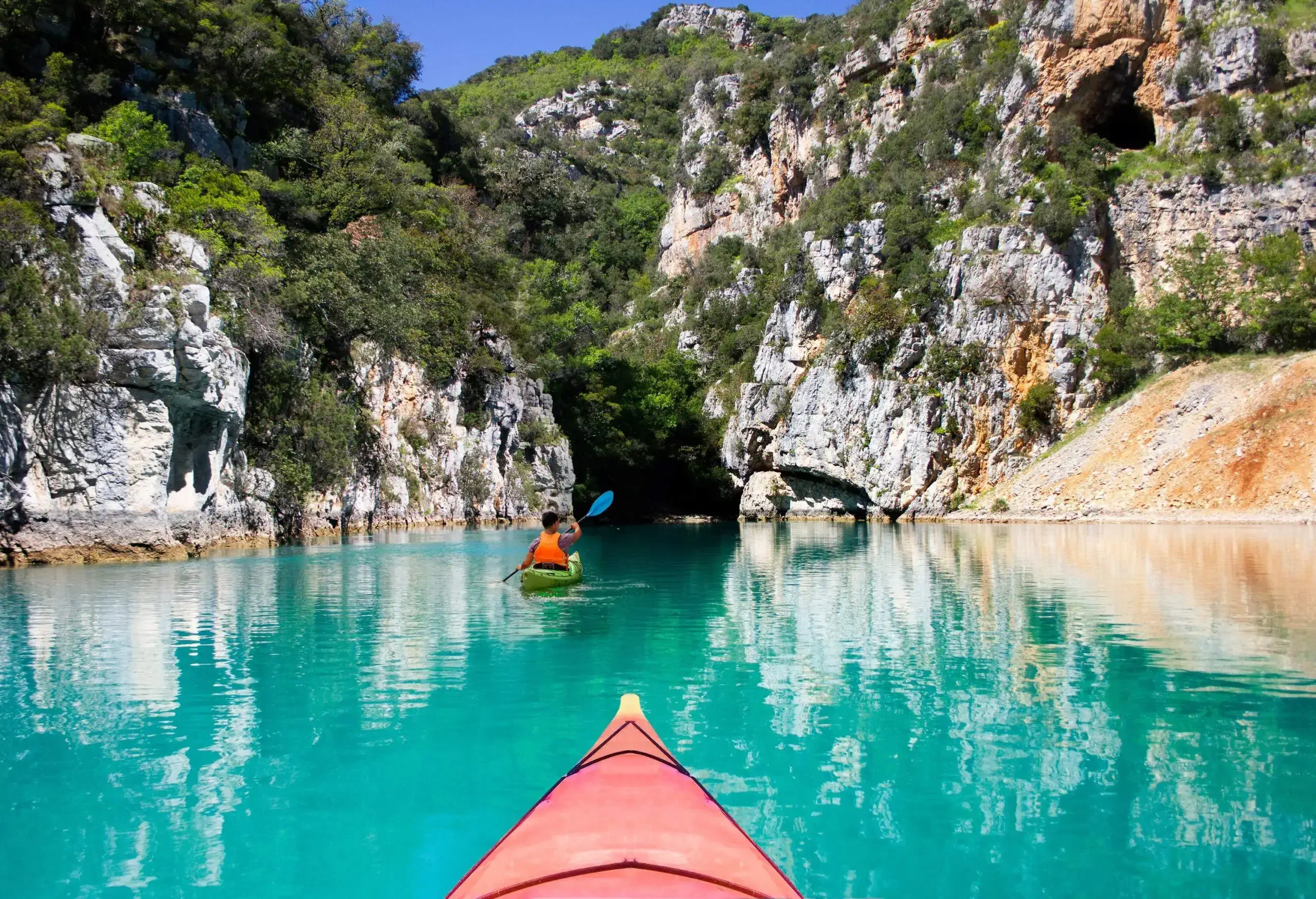A group of kayakers paddles across a tranquil turquoise river in a gorge covered in vegetation.