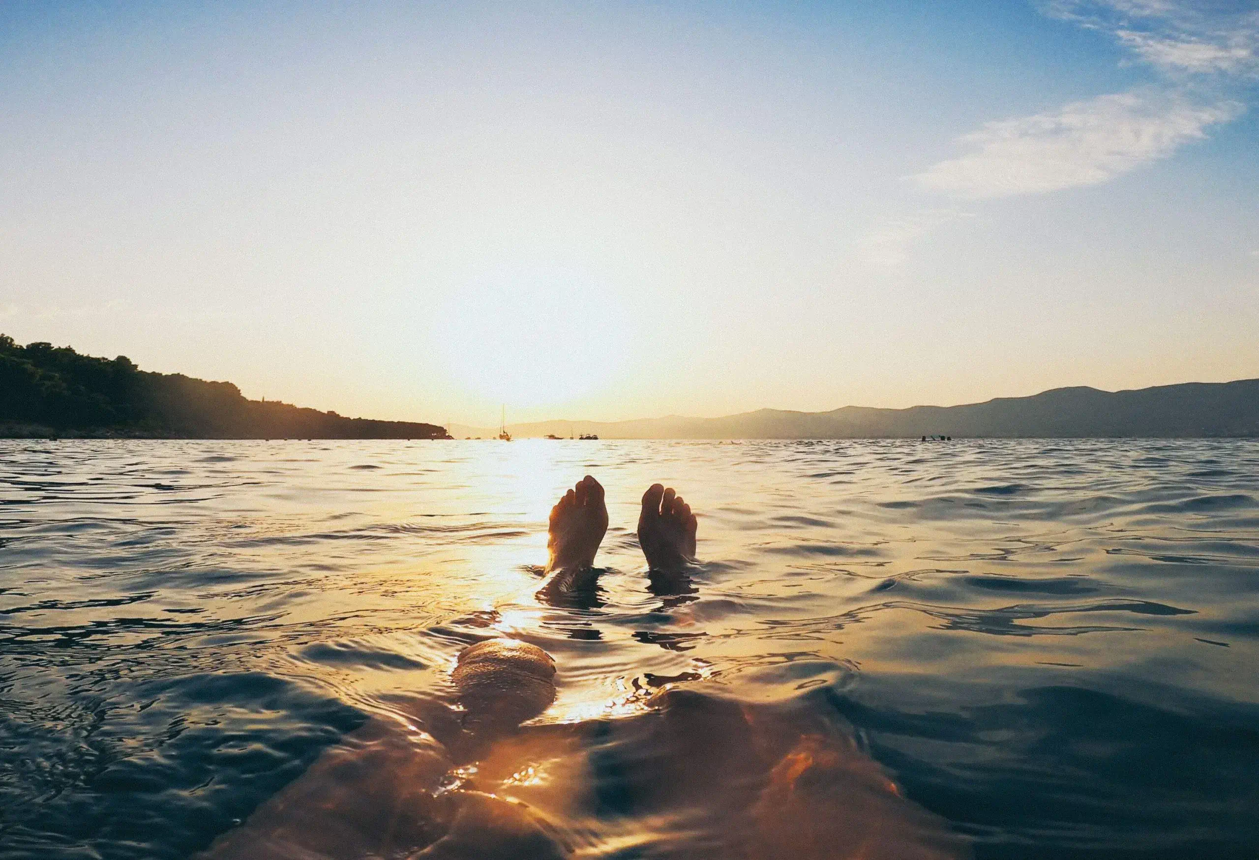 Sunlit bare feet submerged in the water with a view of steep mountain ranges against the bright blue sky.