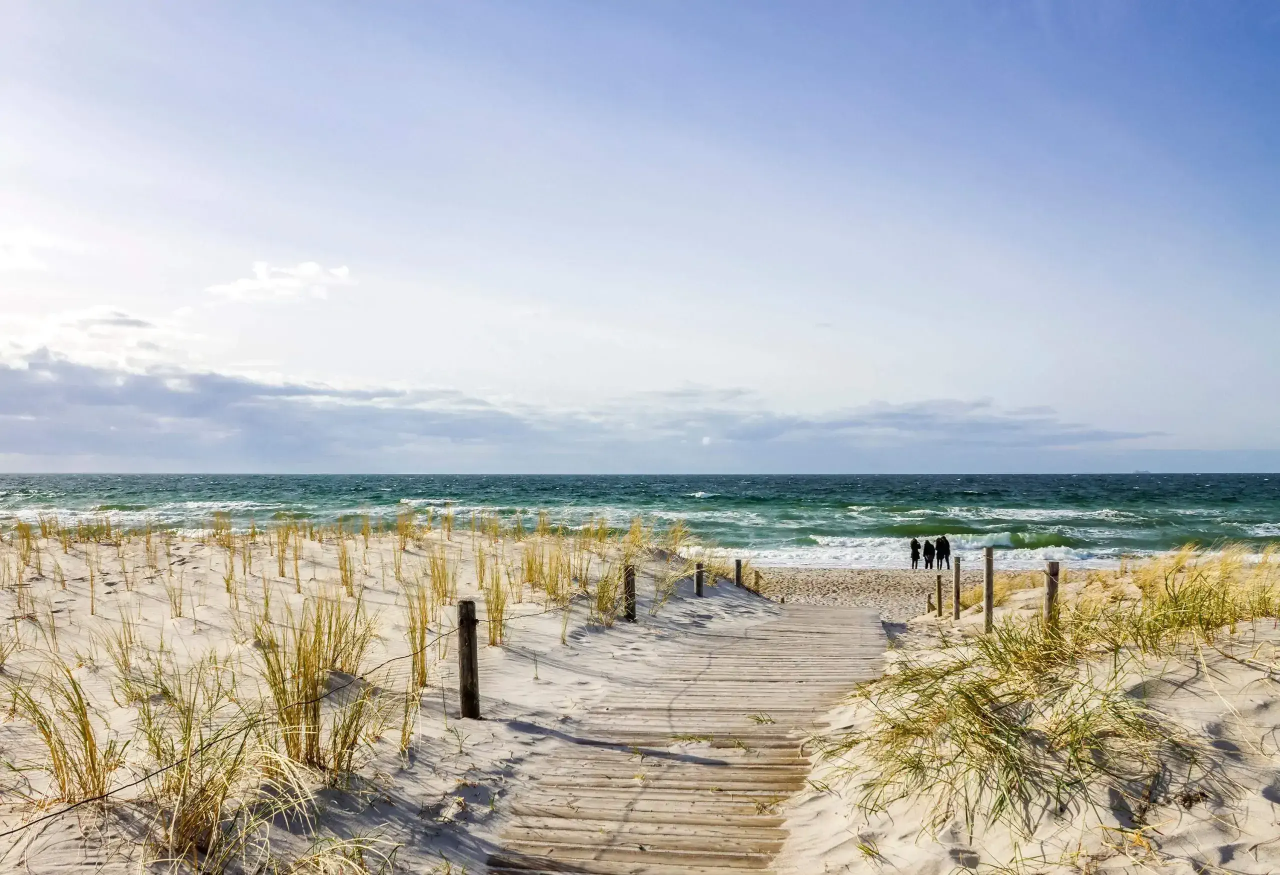 Three silhouetted people stand at the end of a wooden boardwalk by the beach that is surrounded by a wire fence.