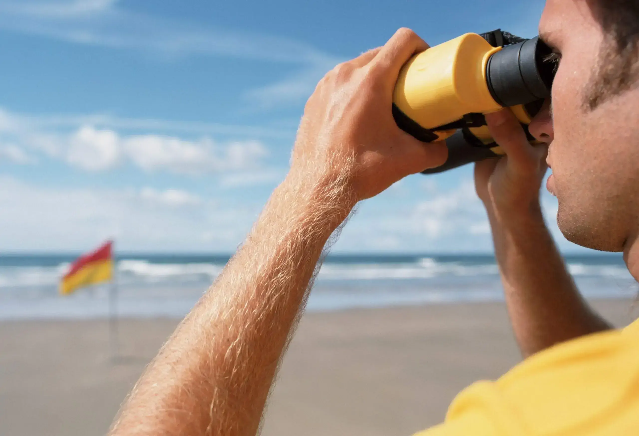 A male lifeguard looking through binoculars at the beach with a red and yellow flag in the background