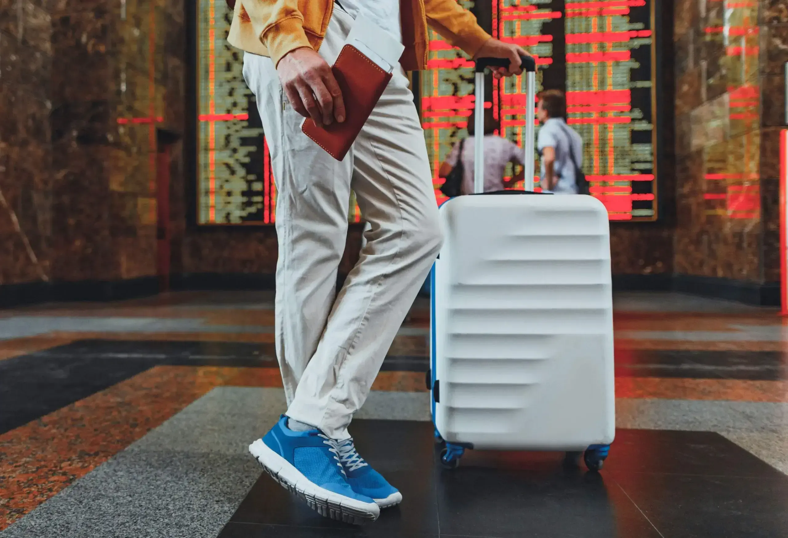 A man wearing blue sneakers standing with one leg over the other next to his white suitcase.