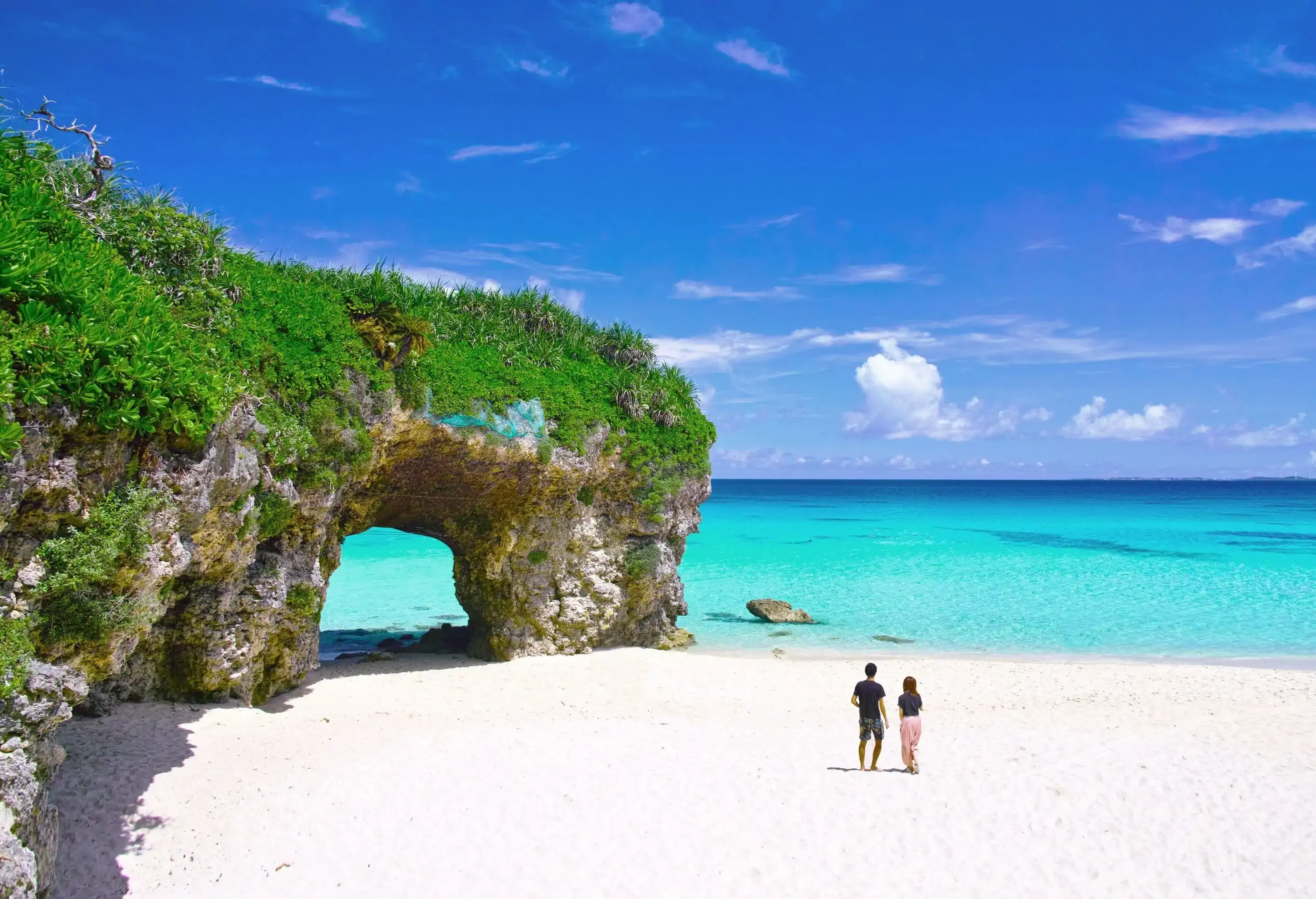 Two people walking across a sandy white beach with a natural arch.
