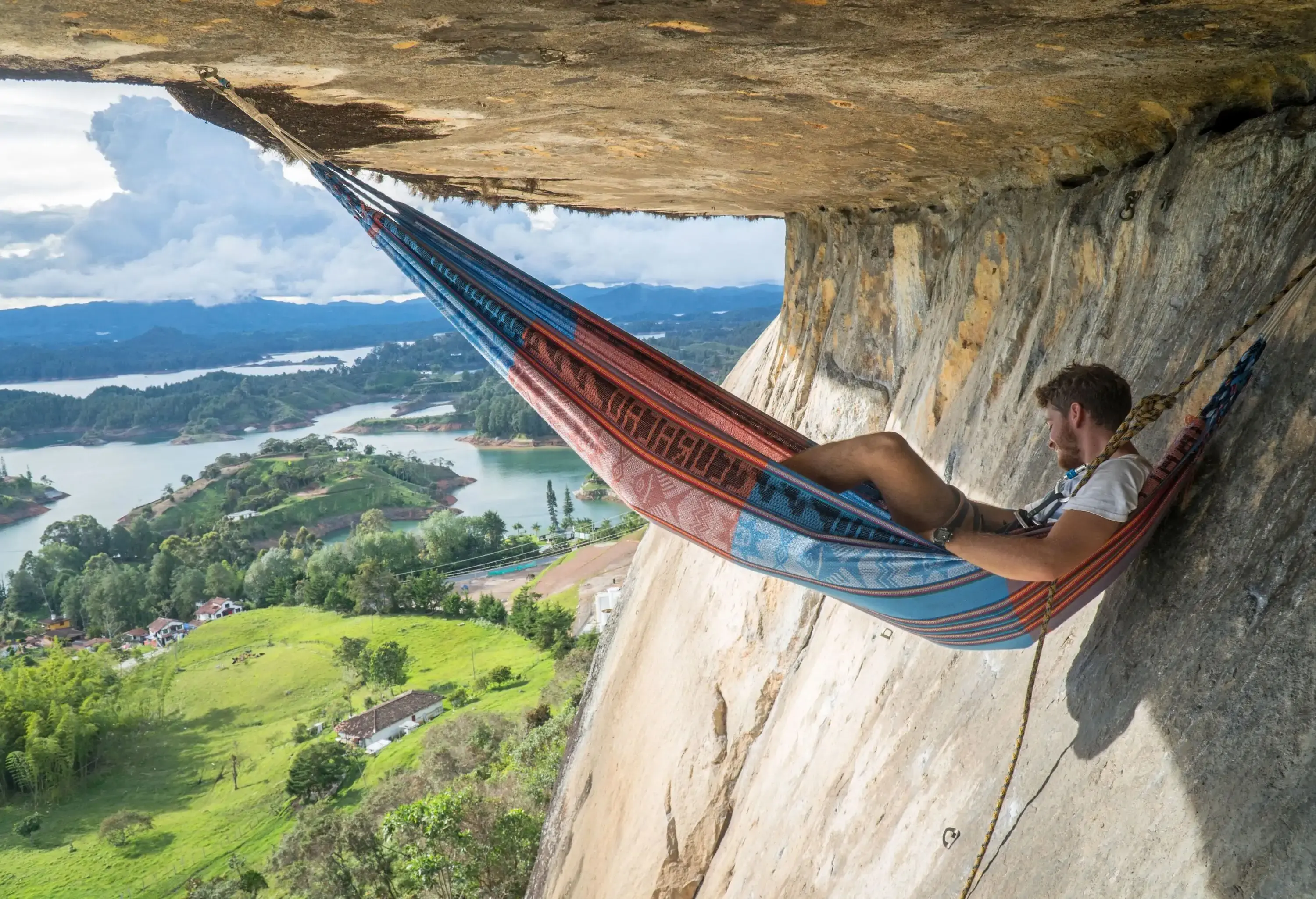 A rock climber relaxes on a hammock hooked to the ceiling of a rock cave overlooking the lake surrounded by small lush islands.