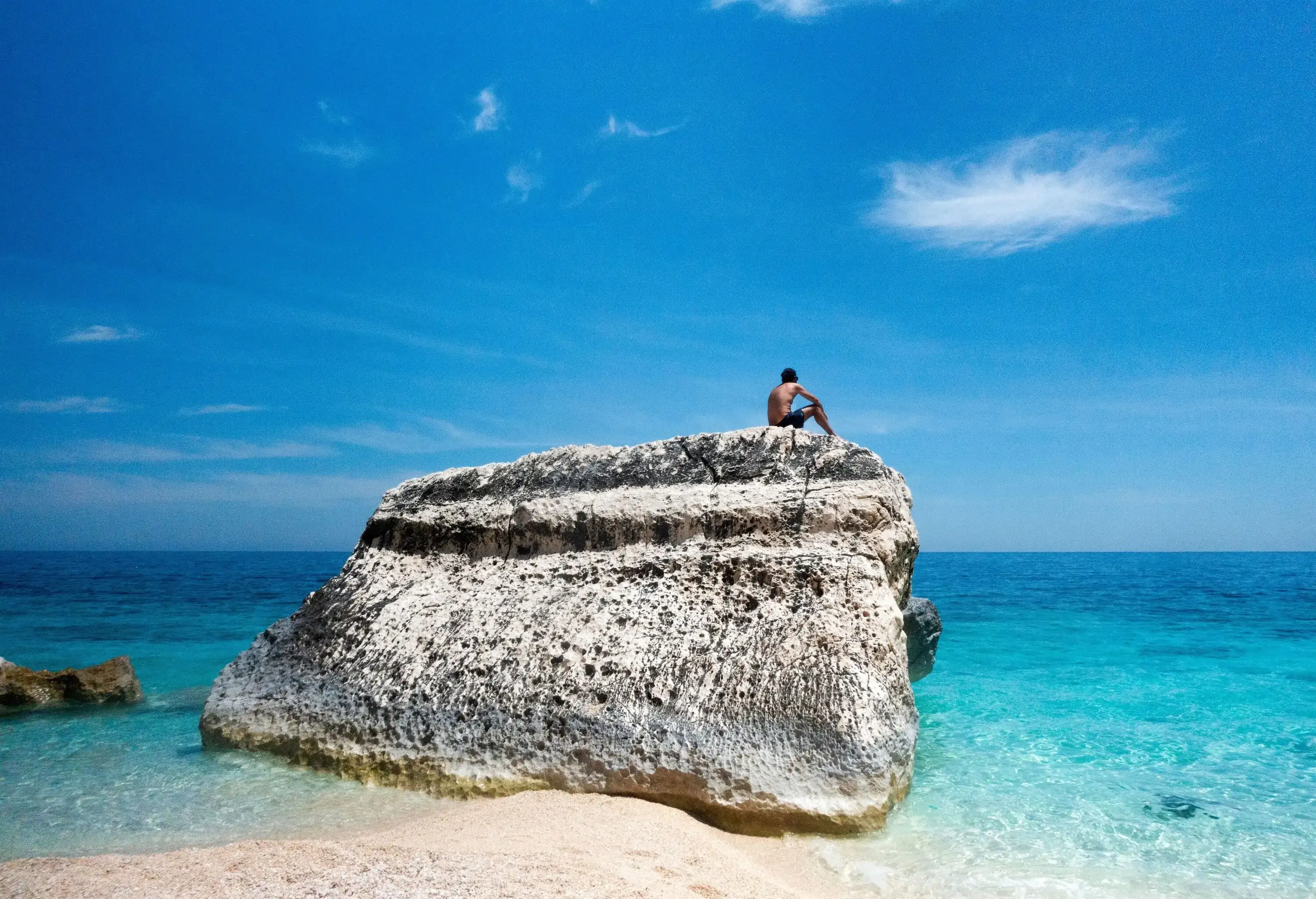 A young man stares at the sea seated on top of a limestone rock, in the seashore of Cala Mariolu, Sardinia in summer