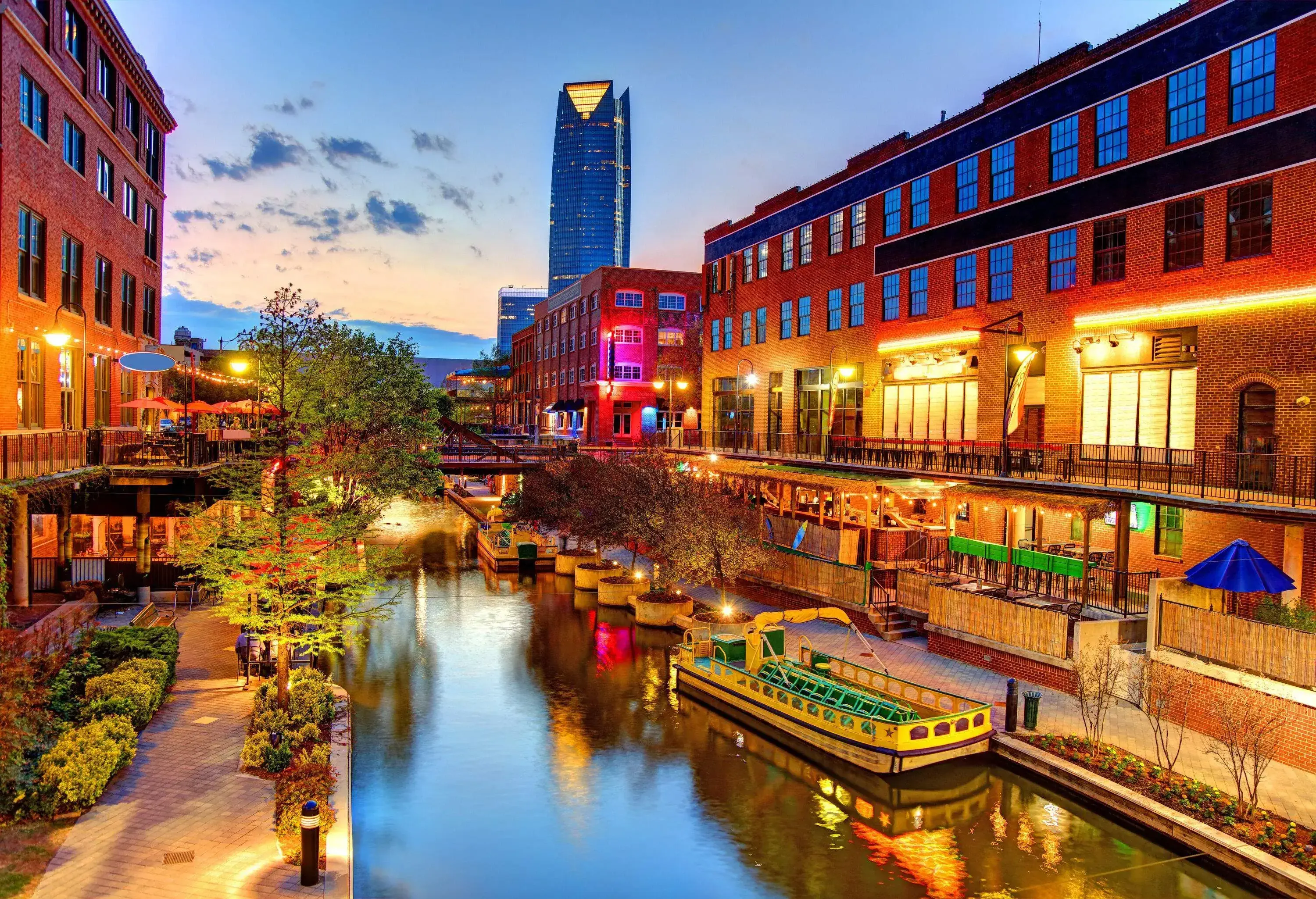 A night-time view of a calm water canal with an anchored tourist boat along a row of trees and commercial establishments.