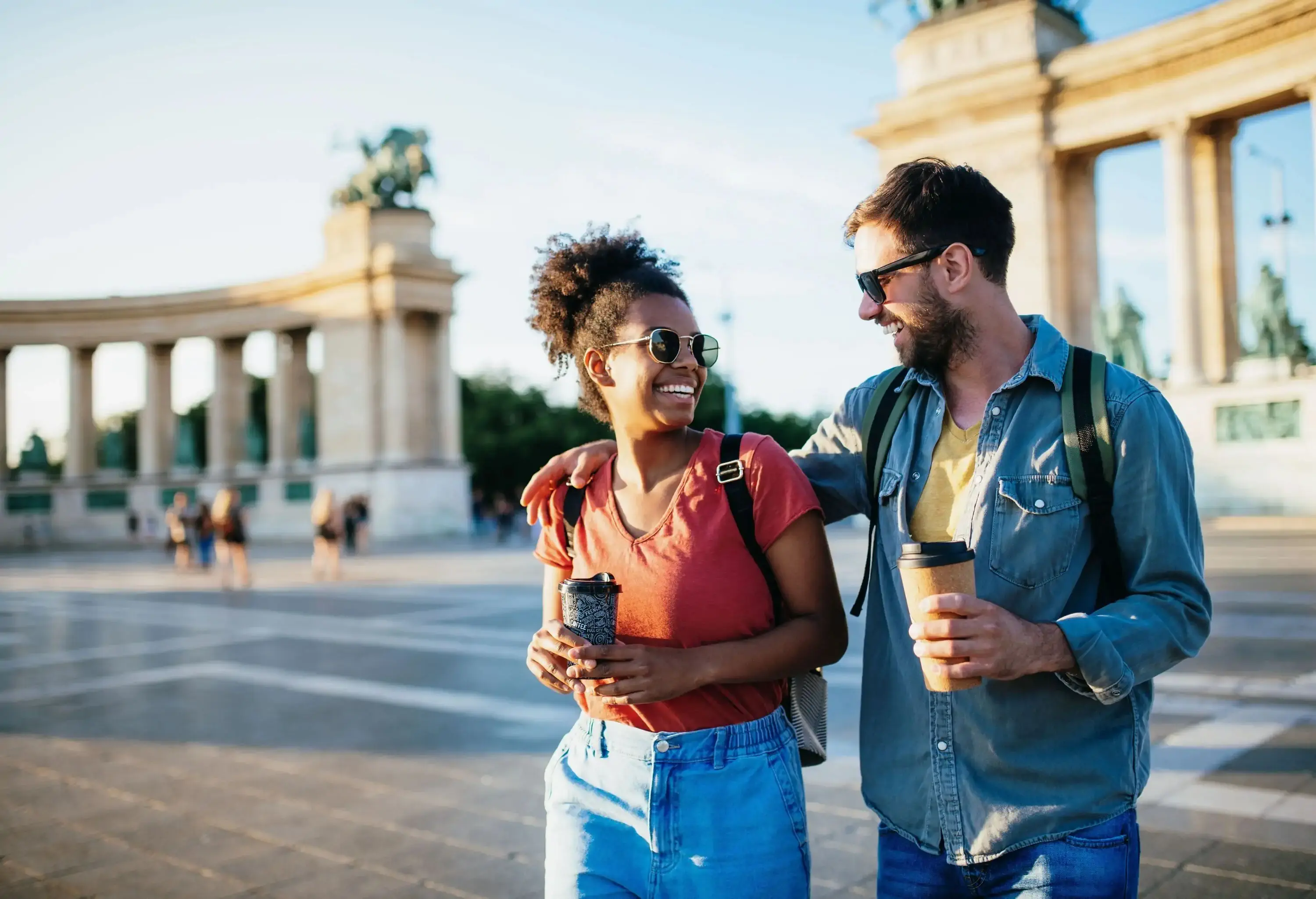 young multiracial couple enjoying city break, holding coffee cups and wearing backpacks
