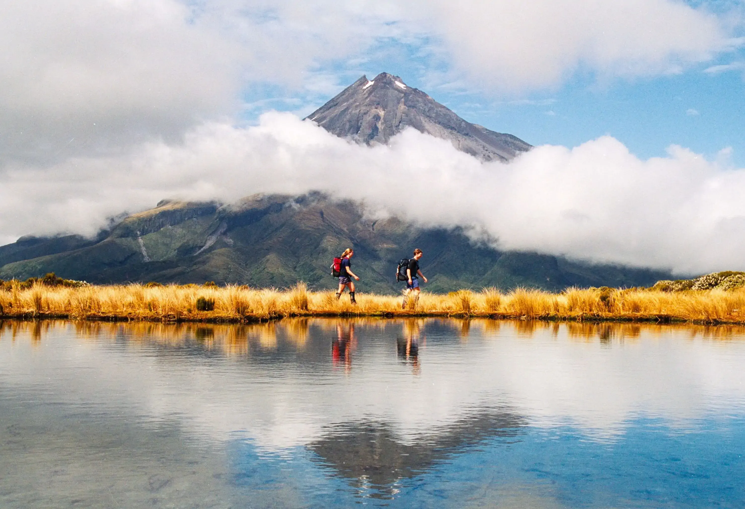 Two people hiking on a grassy area next to a lake with a cloud-covered mountain in the background.