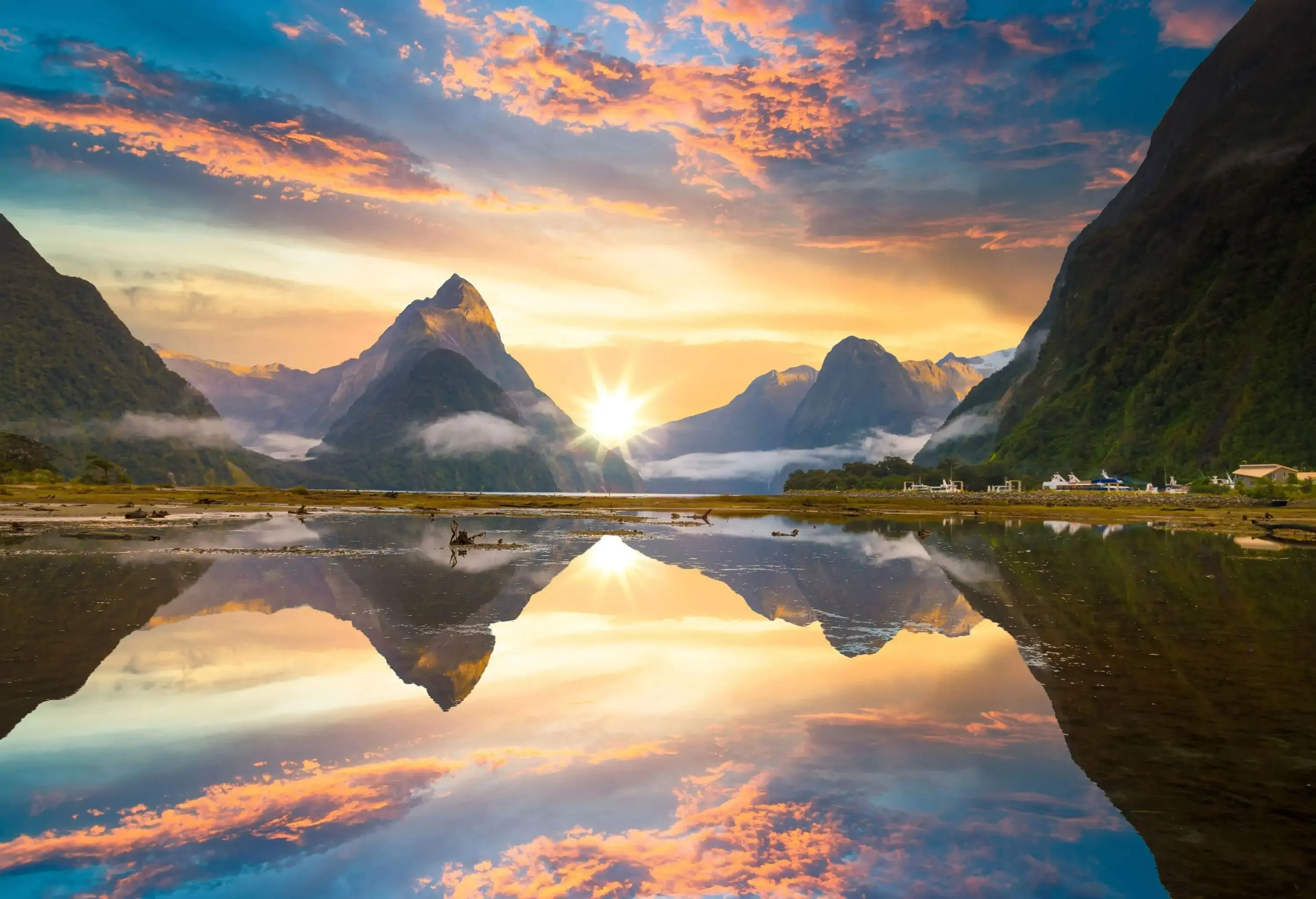 The sun rising over the forested mountain range against the scenic sky mirrors the tranquil water.