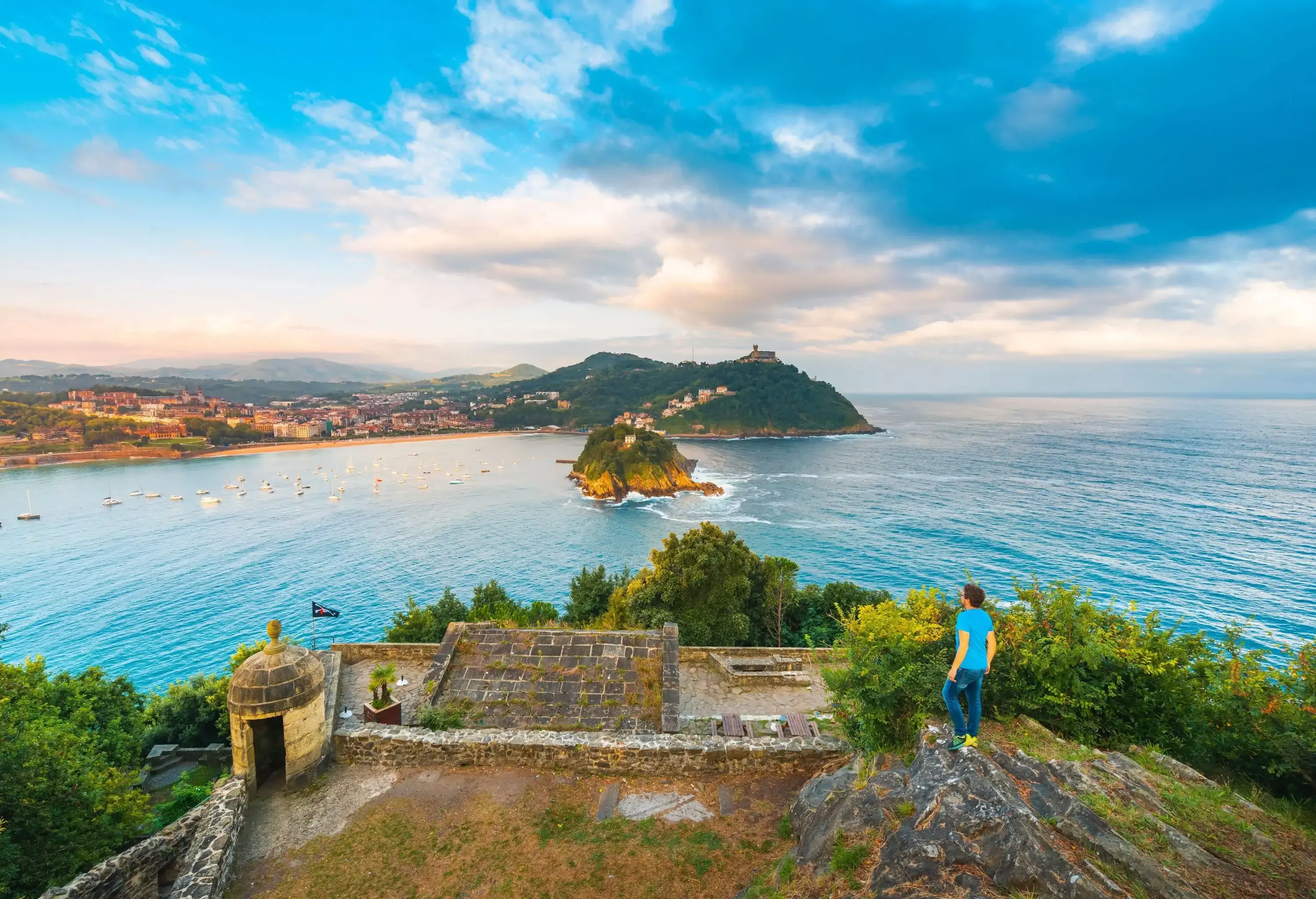 A person stands on a cliff's edge with a brick platform overlooking an island in a bay with cruising boats.