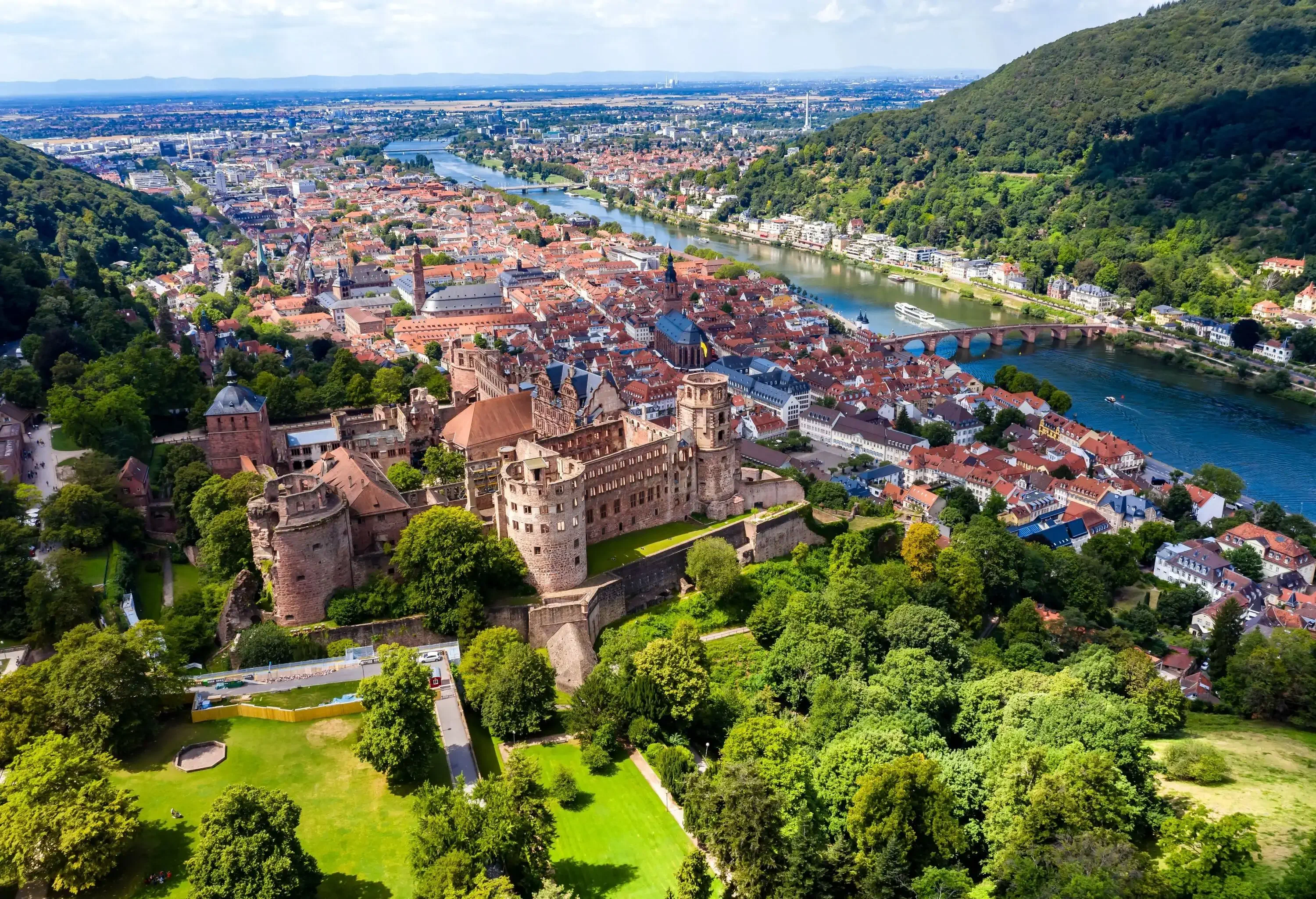 Aerial view of an old city by the river surrounded by forested green hills and a ruined sandstone castle in the centre.