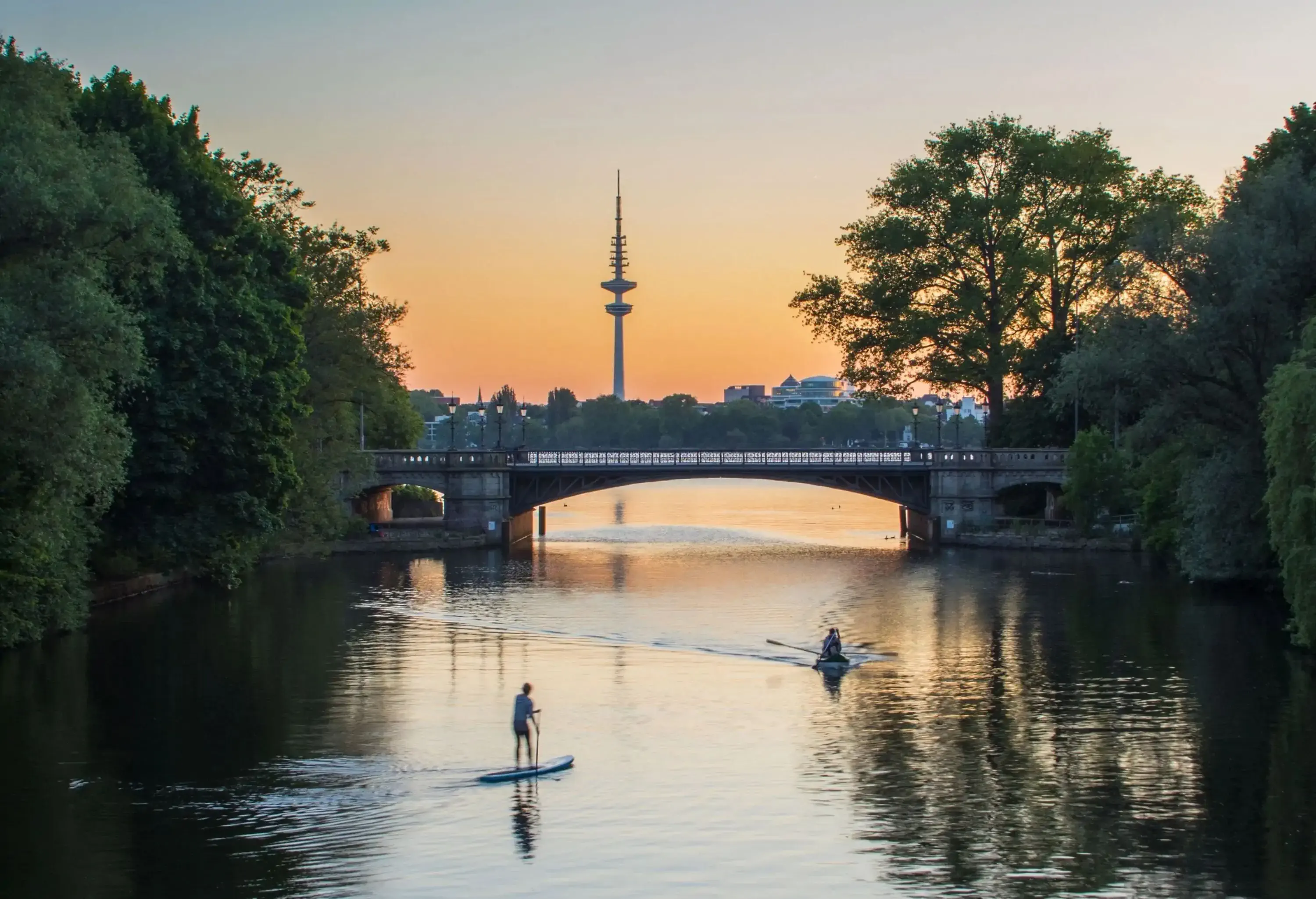 A bridge across a calm lake with people paddleboarding and kayaking under the sunset skies.