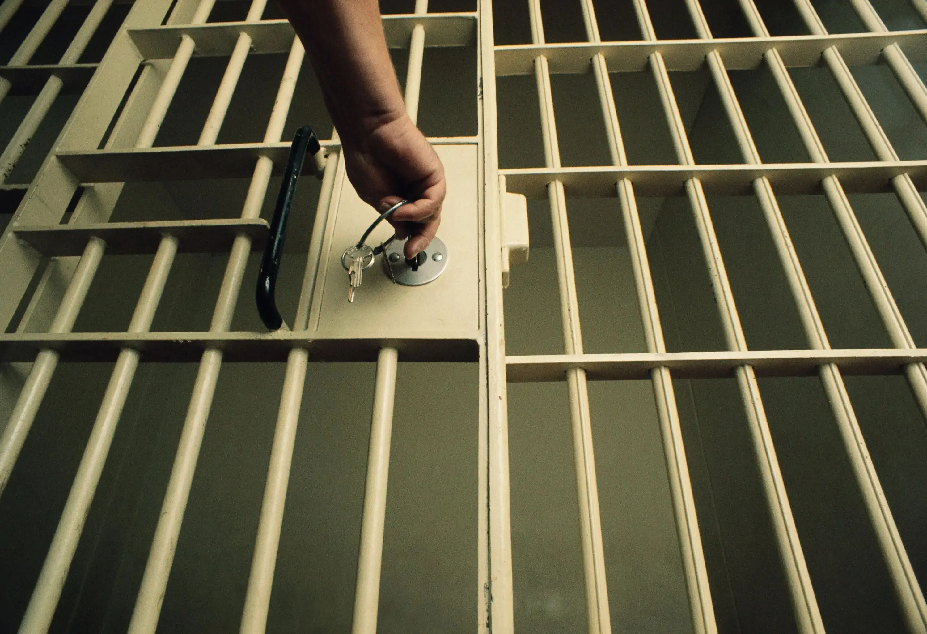 A hand opens a prison cell door of an empty room.
