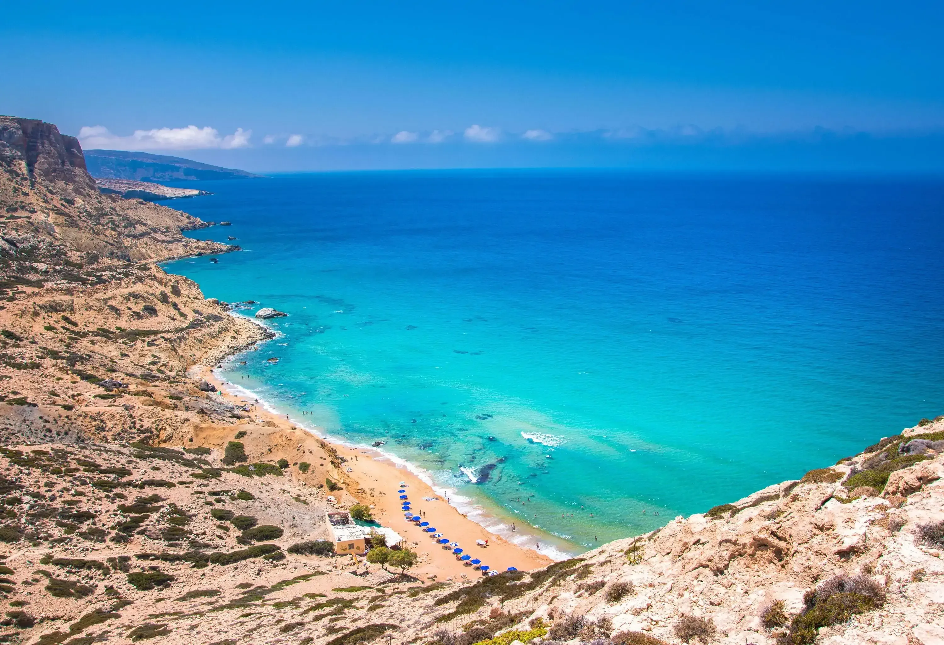 A rocky outcrop is next to a narrow beach with parasols and sun loungers that all face the huge expanse of blue sea. 