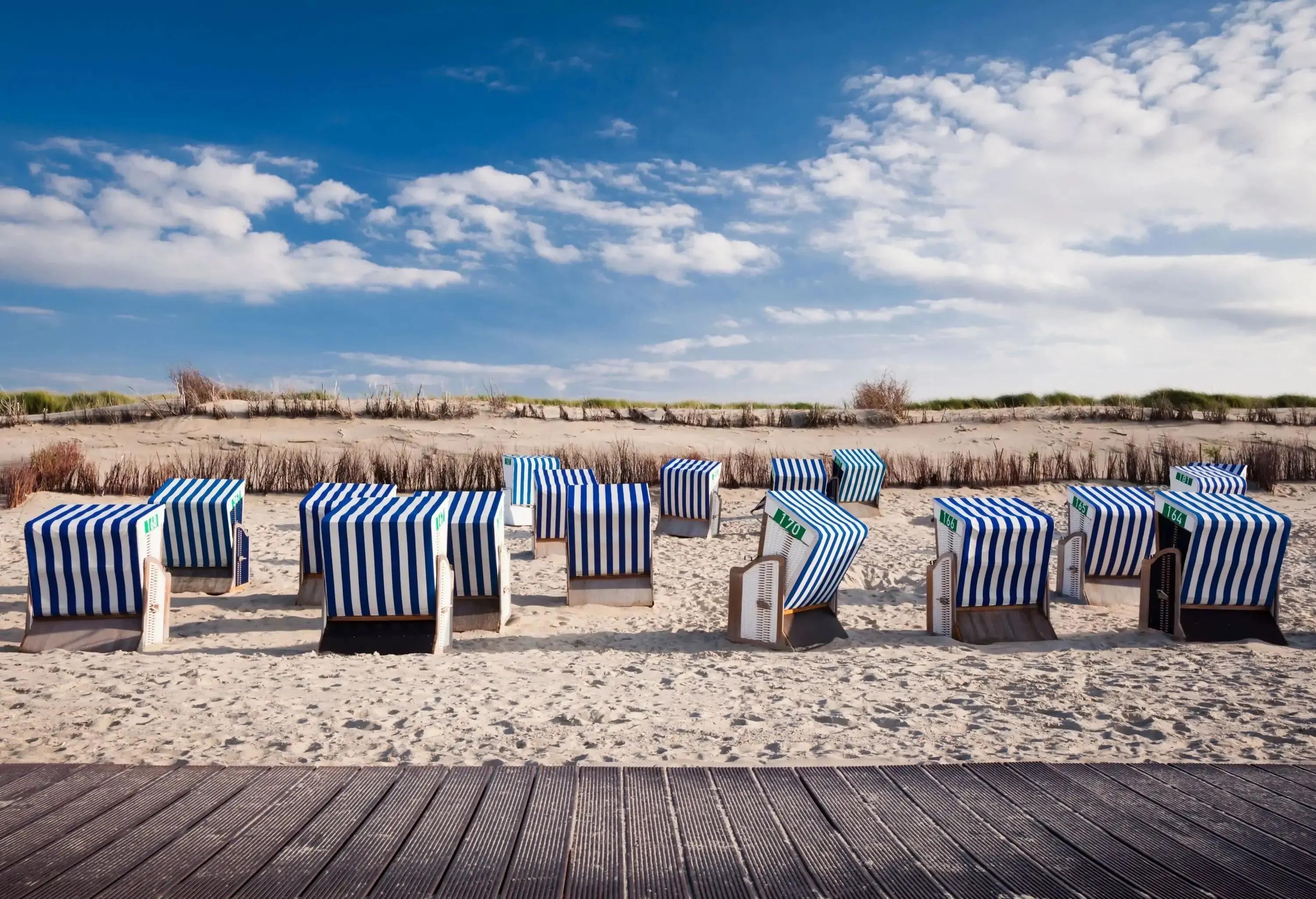 Several blue and white striped hooded beach baskets on bushy sand alongside a boarded path.