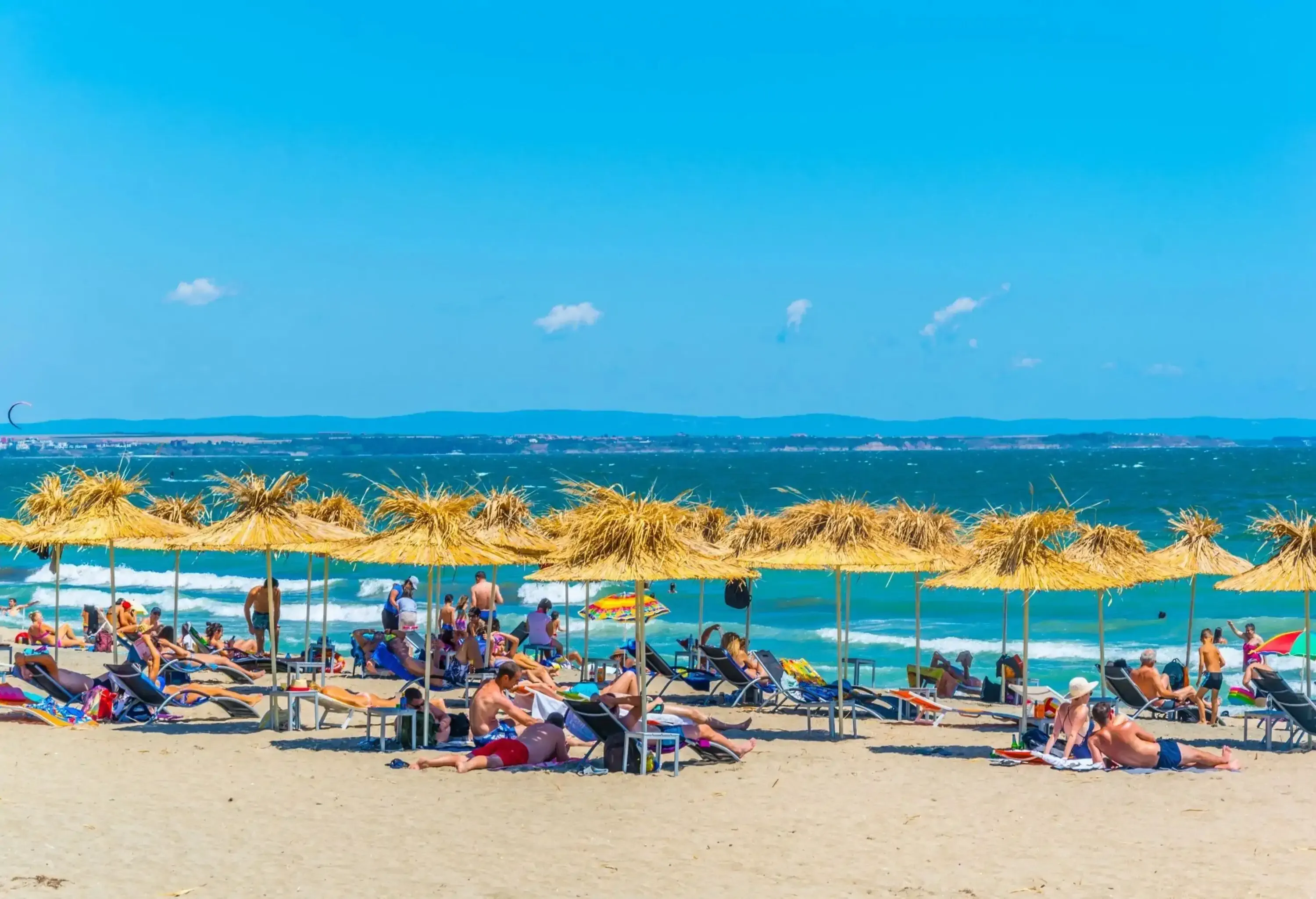 People enjoy the bright sun as they bask on the shore of a serene sea, with sun loungers and straw parasols lining the beach.