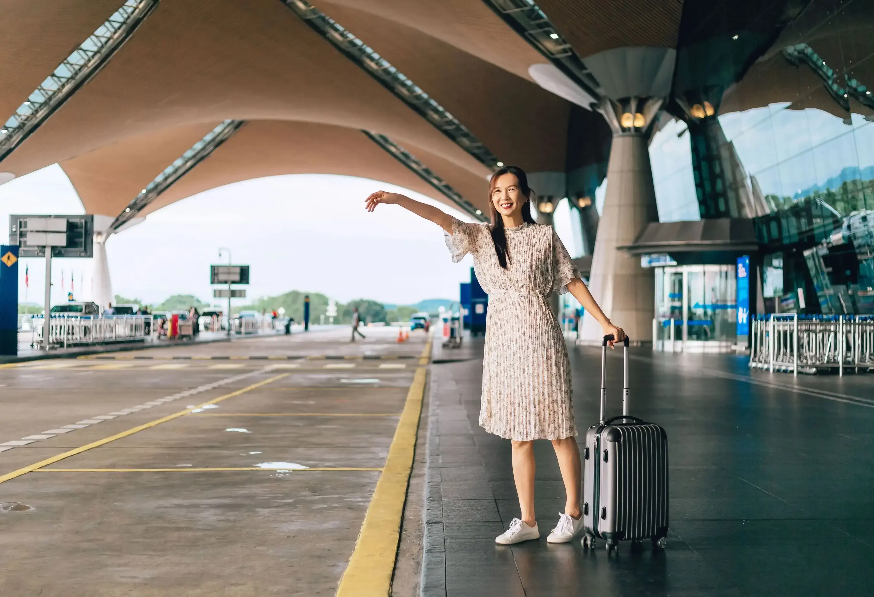 A smiling woman standing at a modern airport curb, holding a suitcase while gesturing with her arm.