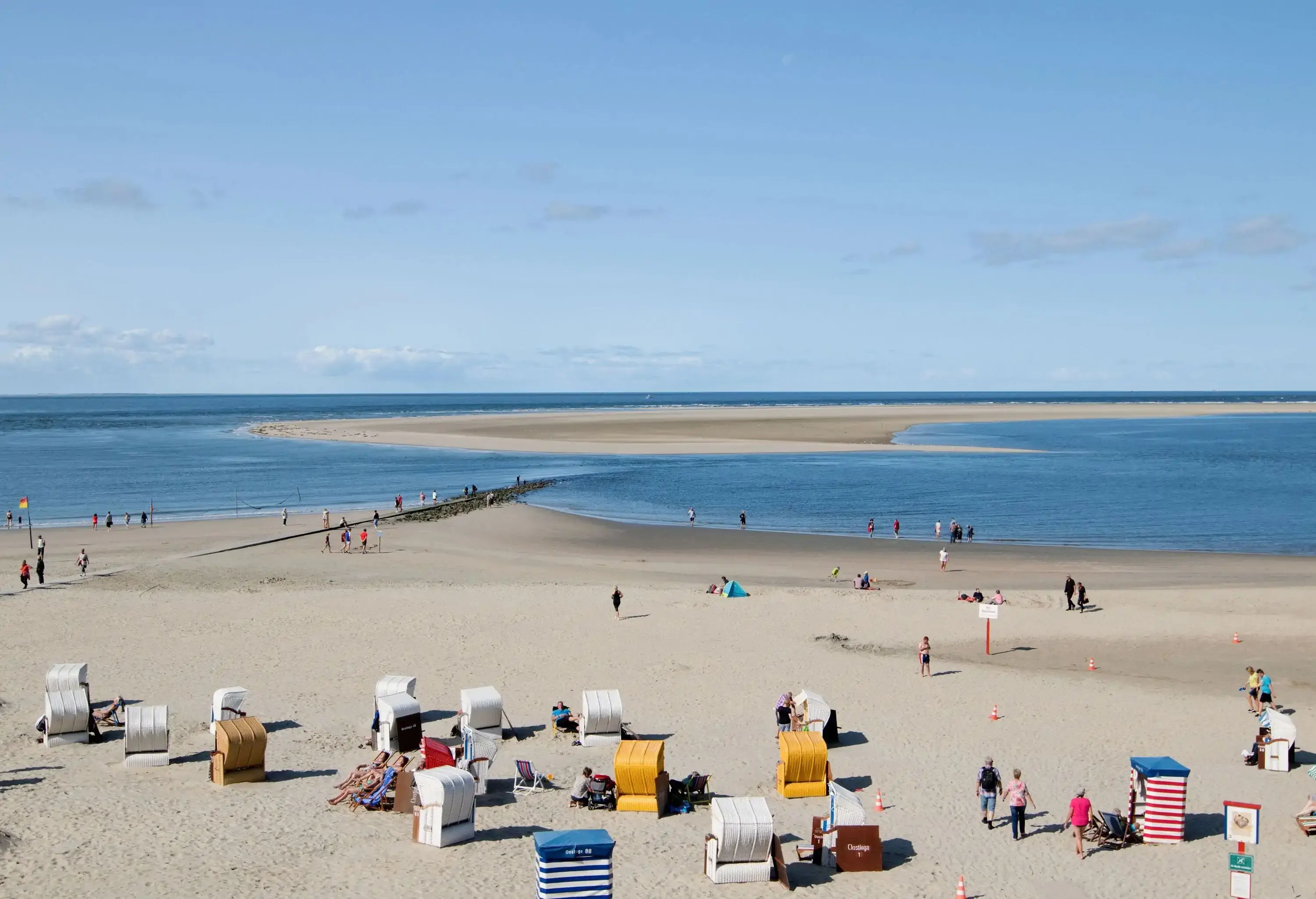 A number of wicker chairs with canopies are placed in the sand close to the water's edge, where beachgoers lounge and swim.