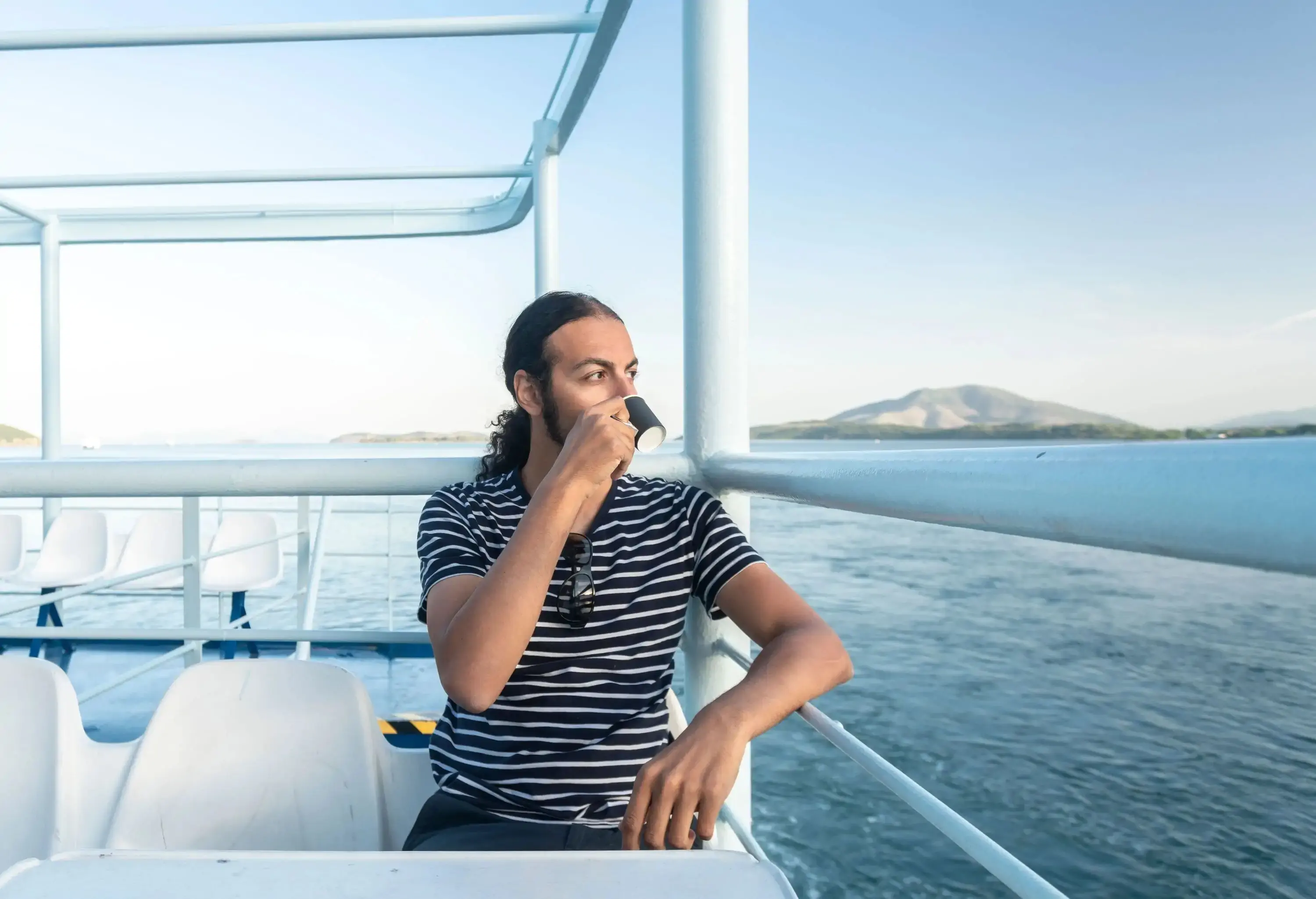 A man in a striped shirt sits on a passenger boat while drinking coffee.