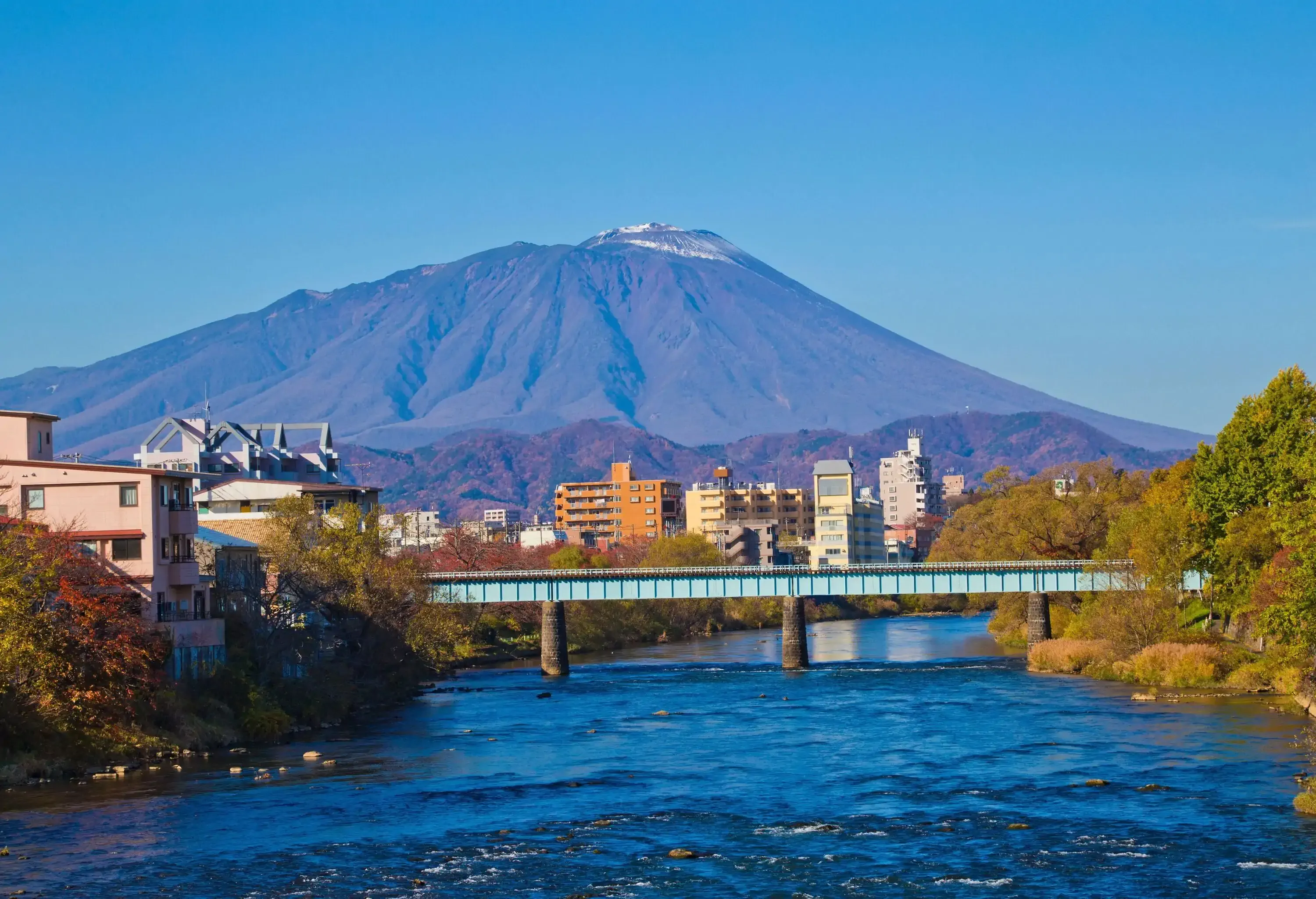 A picturesque mountain in contrast with the blue sky in the backdrop, a bridge span the dazzling river amidst the lush trees.