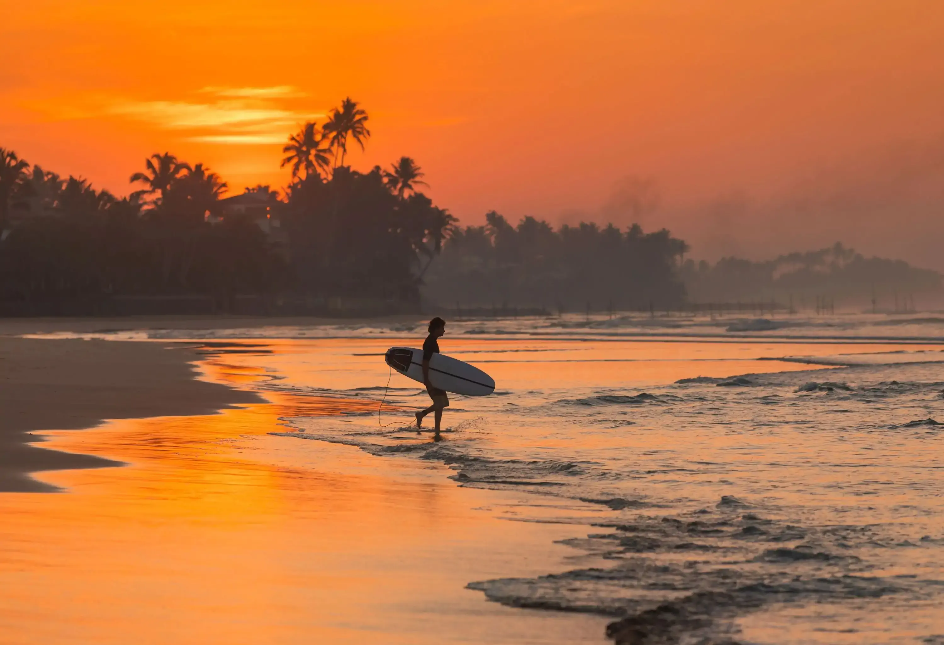 A man walking across the beach towards the sea with a surfboard on his side.