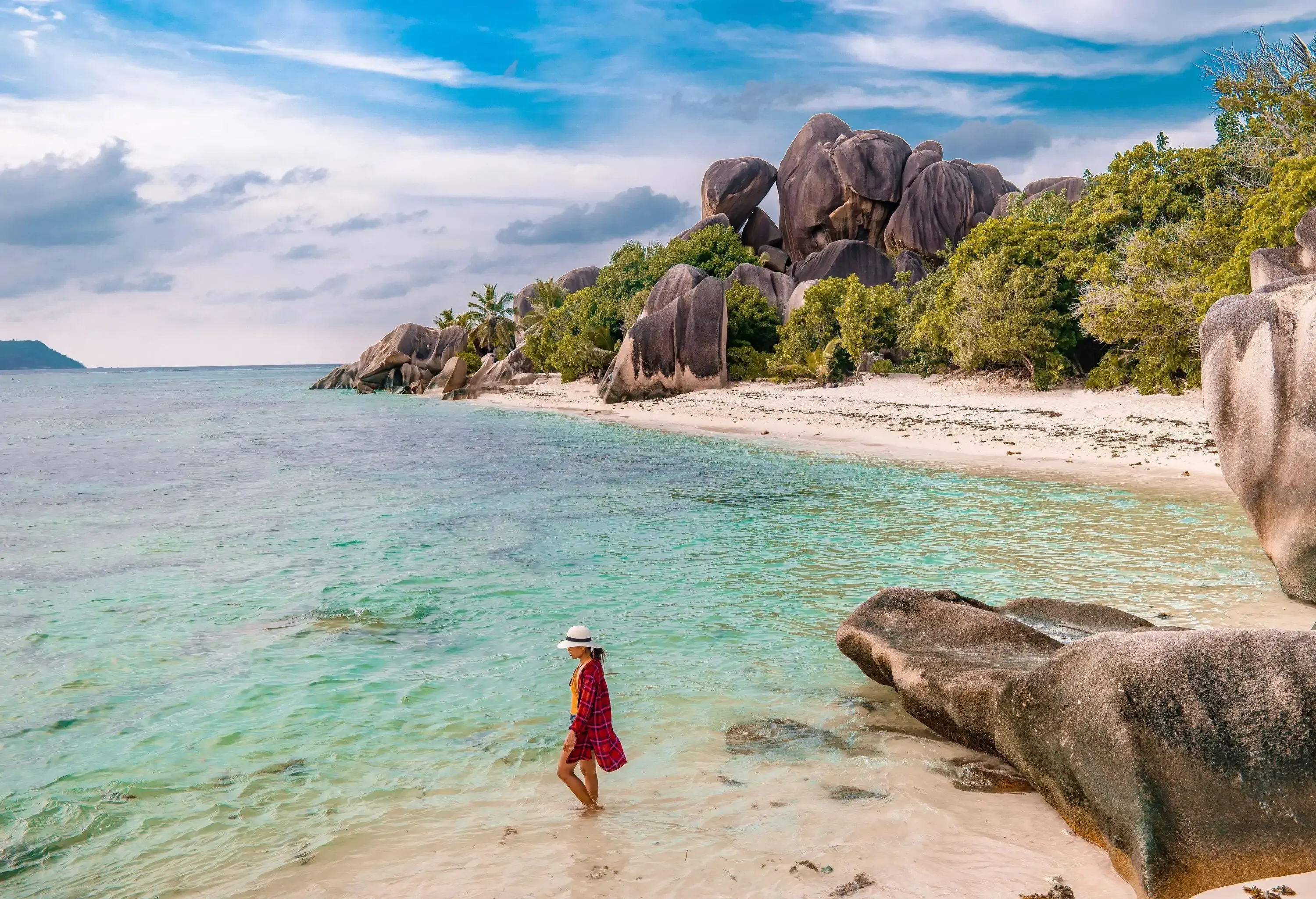A woman in a chequered shirt and hat stands in the crystal-clear waters of a beach along big boulders and lush vegetation.