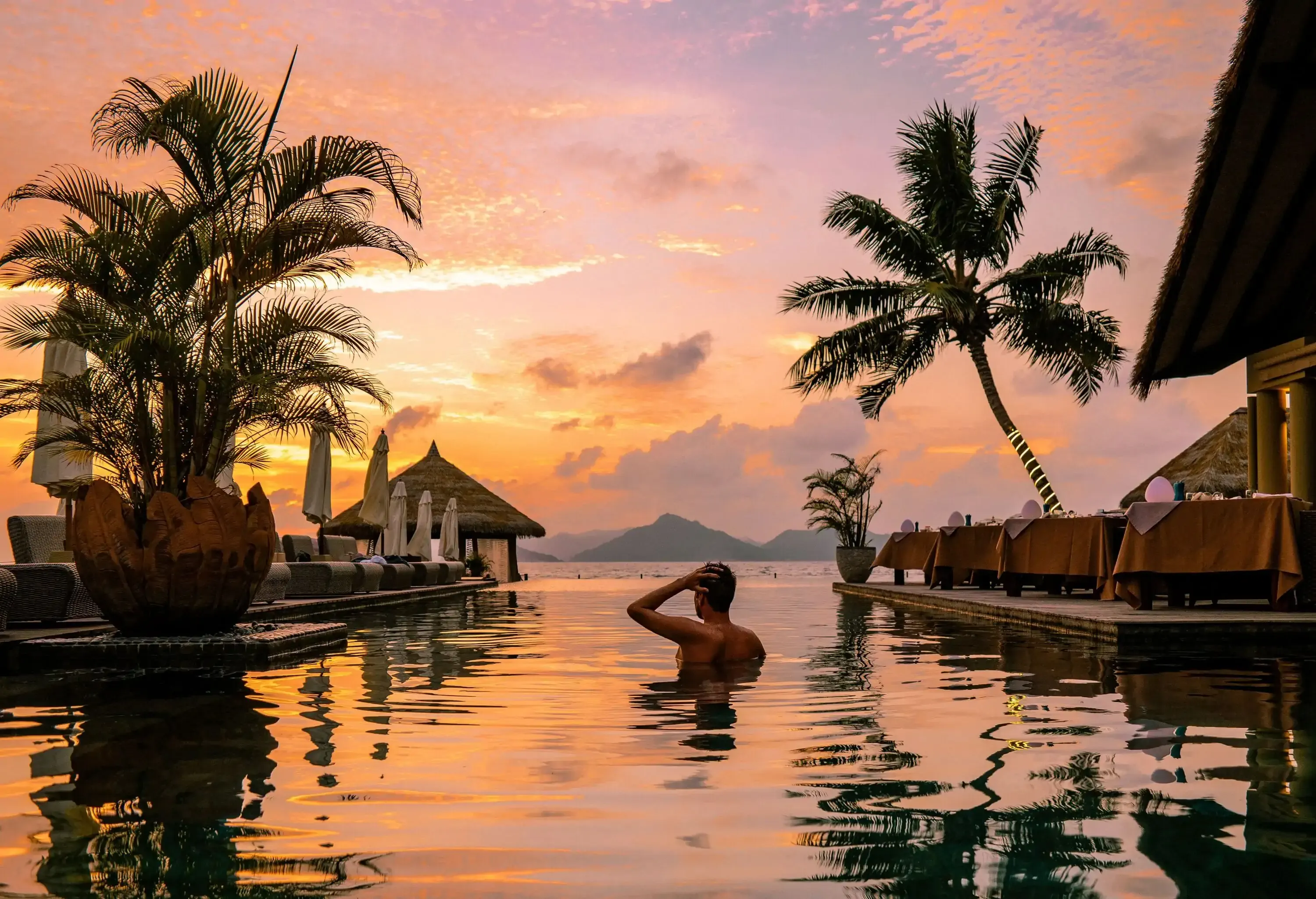 A person in an infinity pool at sunset, framed by silhouetted palm trees and resort structures against a colorful sky.