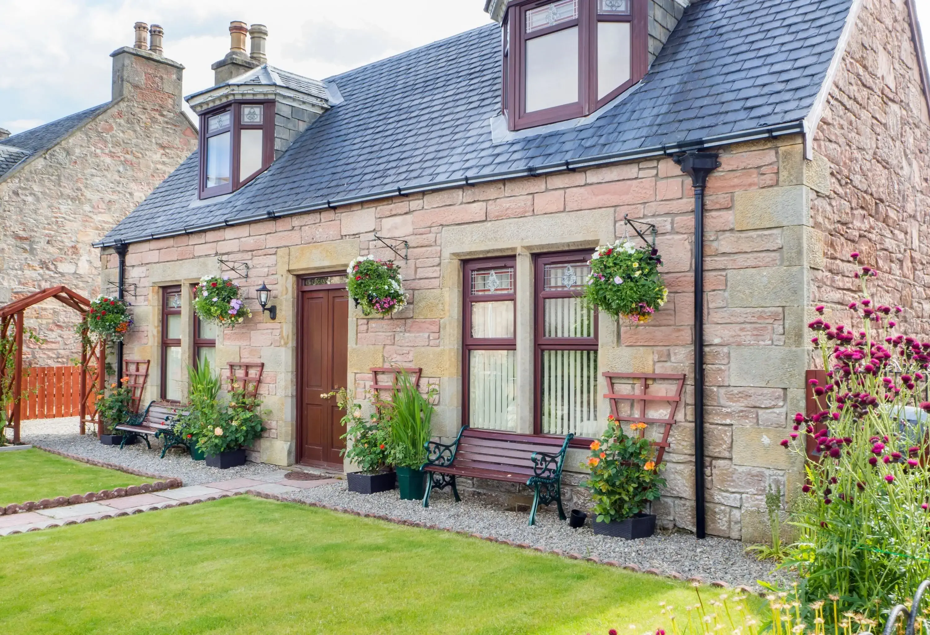 A stone cottage house with outdoor benches, flower planters, dormer windows, and raised chimneys.