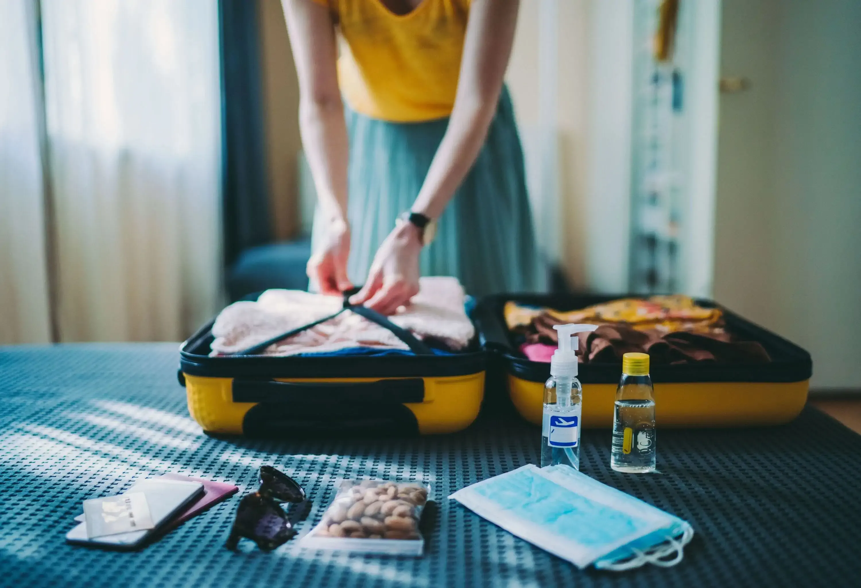 A traveller packing clothes into the yellow suitcase and preparing other kinds of stuff like a passport, alcohol, and facemasks for her hand carry.