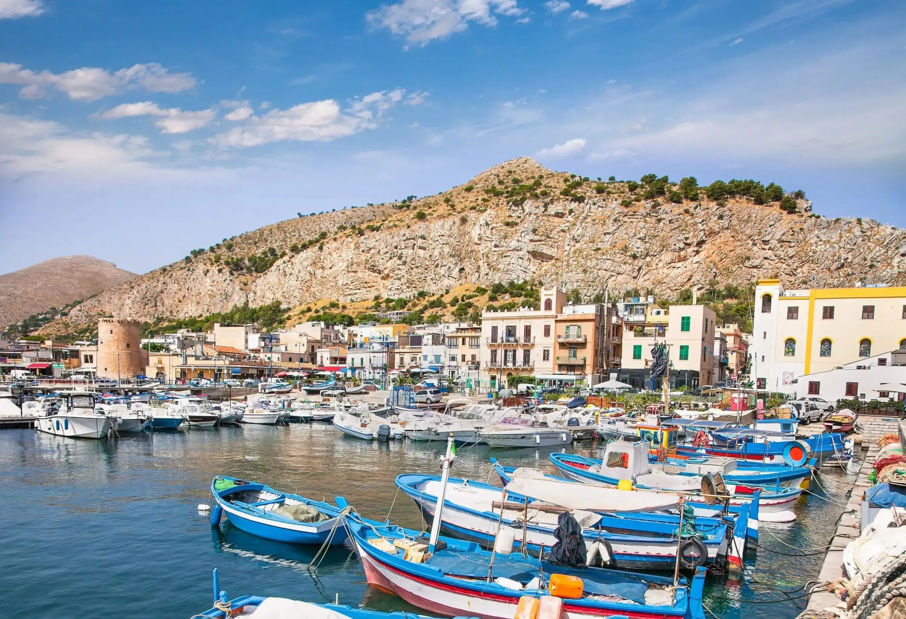 Colourful boats docked at a port lined with buildings, with a rugged hill in the background, against a backdrop of blue sky and sea.
